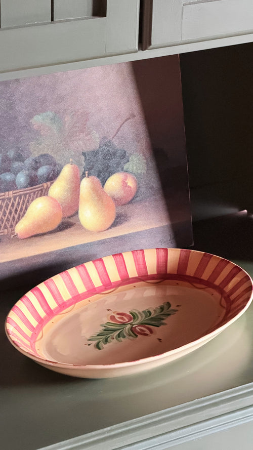 A Vintage Gail Pittman Siena Serving Platter displayed on a shelf in front of a still life fruit painting.
