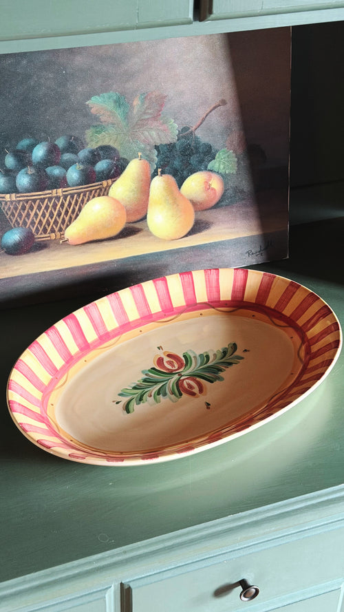 An oval ceramic serving platter with red and yellow stripes and a green leaf motif, displayed on a green dresser in front of a framed still life painting.