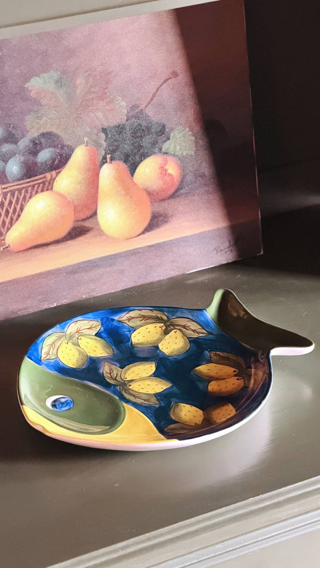 A Vintage Studio Nova Lemon Fish Serving Platter with a painted yellow and blue pattern, displayed on a shelf in front of a still life fruit painting.