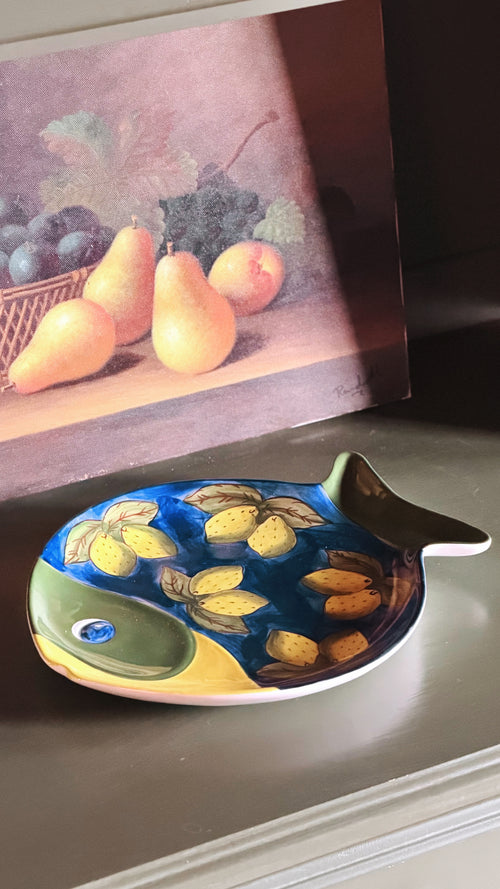 A Vintage Studio Nova Lemon Fish Serving Platter with a painted yellow and blue pattern, displayed on a shelf in front of a still life fruit painting.
