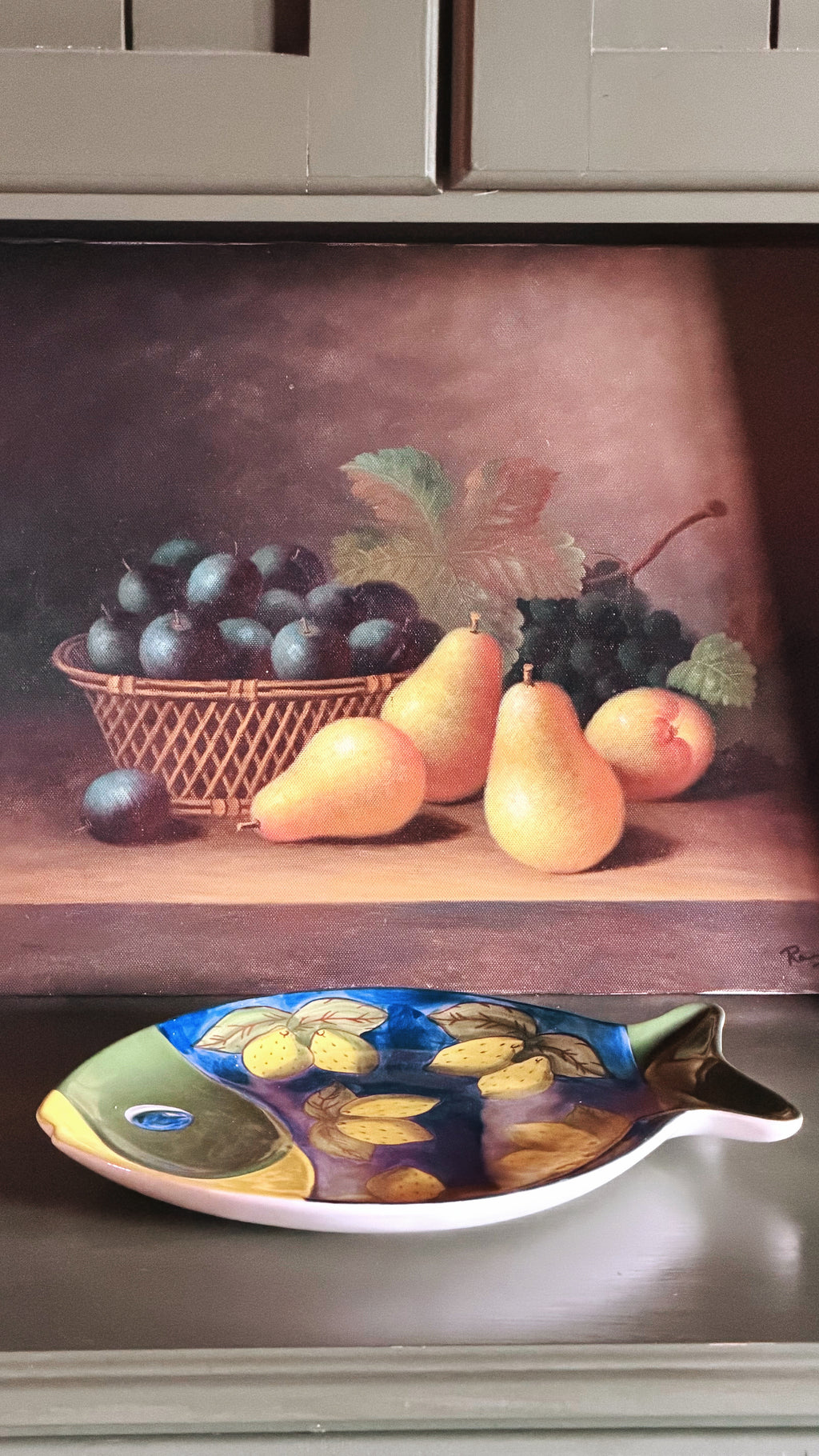 A Vintage Studio Nova Lemon Fish Serving Platter, displayed on a shelf in front of a framed still life painting of grapes and pears.