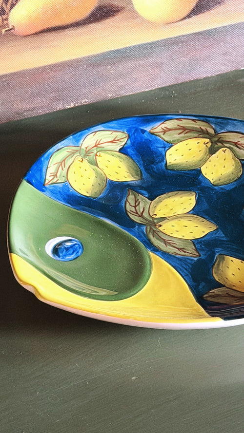 A hand-painted ceramic fish-shaped dish with yellow lemons on a blue background, featuring a green and yellow lower section, shown on a dark green surface.