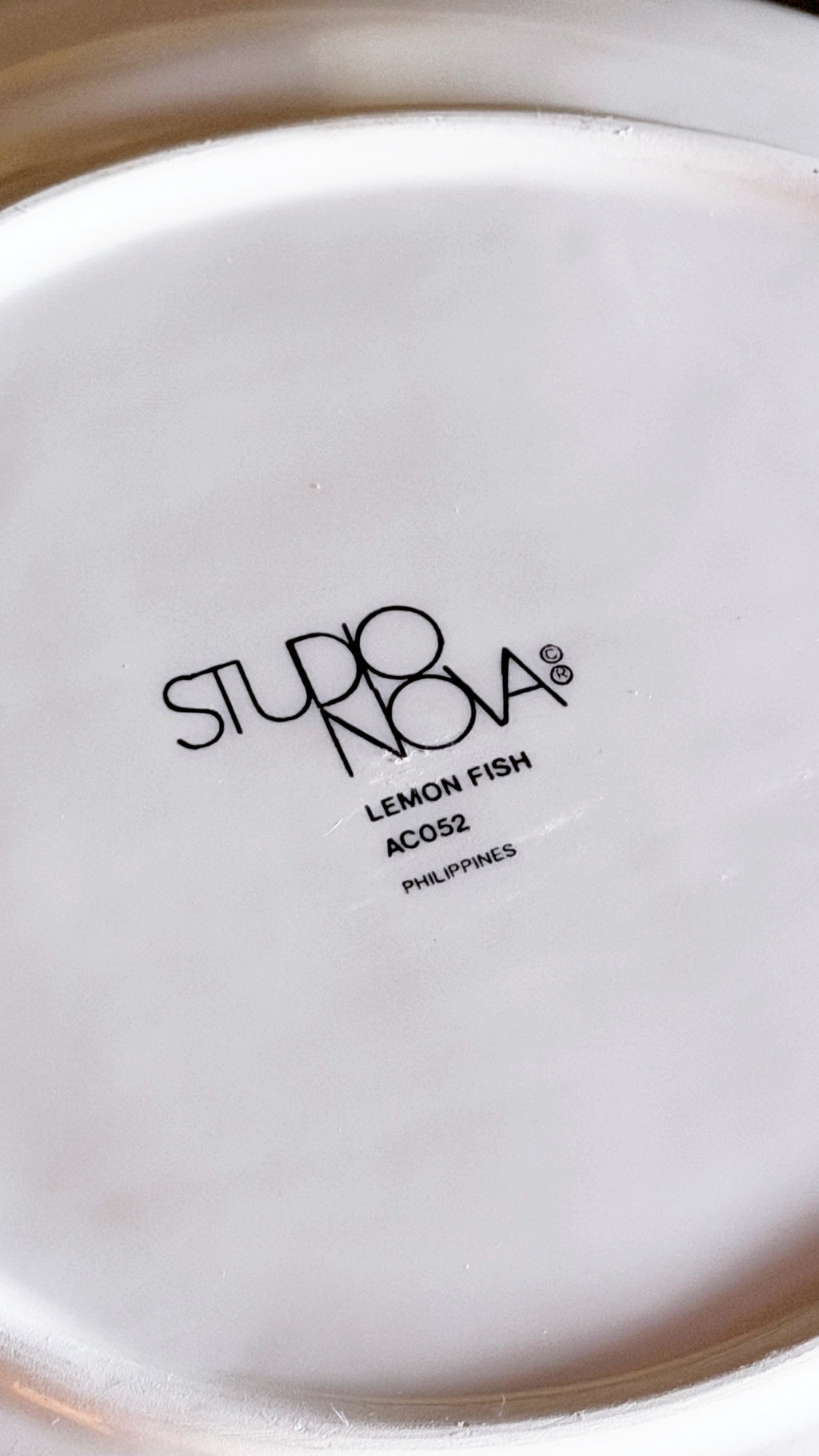 Close-up of the bottom of a white ceramic plate or dish with black printed text reading 'STUDIO NOVA', 'LEMON FISH', 'AC052', and 'PHILIPPINES'.