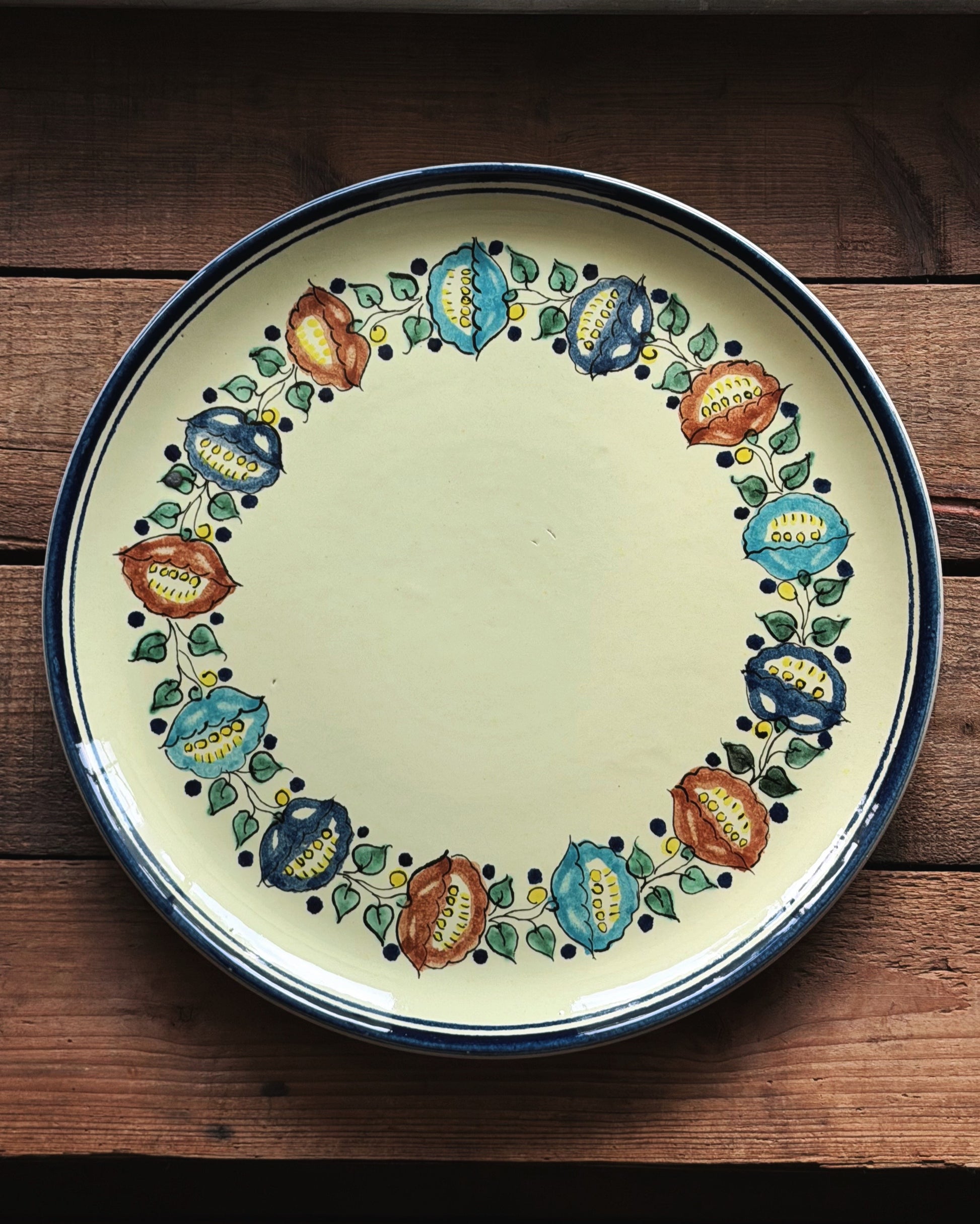 A round ceramic serving platter with a yellow center and a hand-painted floral border in blue, green, red, and yellow, set on a rustic dark wood background.