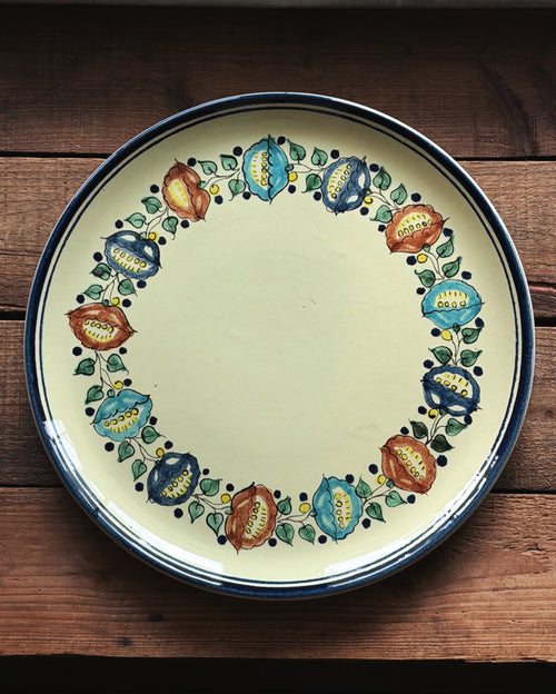 A round ceramic serving platter with a yellow center and a hand-painted floral border in blue, green, red, and yellow, set on a rustic dark wood background.