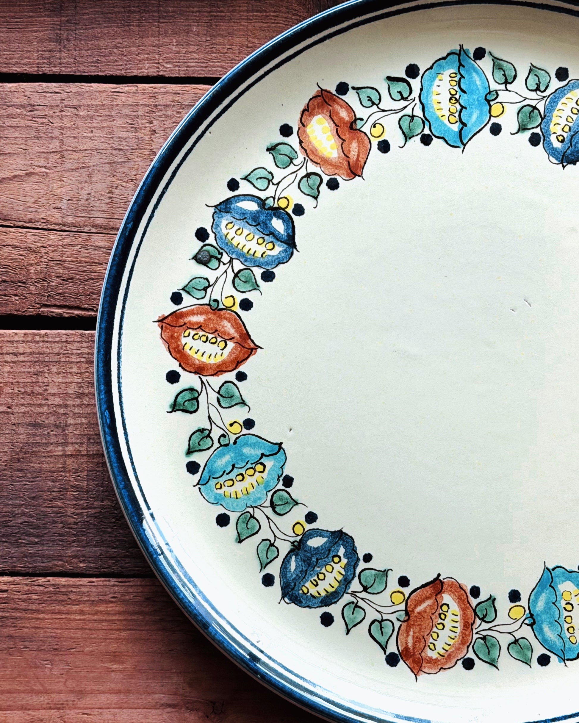 A close-up of a ceramic platter with a hand-painted floral folk art border in blue, orange, and green on an off-white background, set on a rustic dark wood surface.
