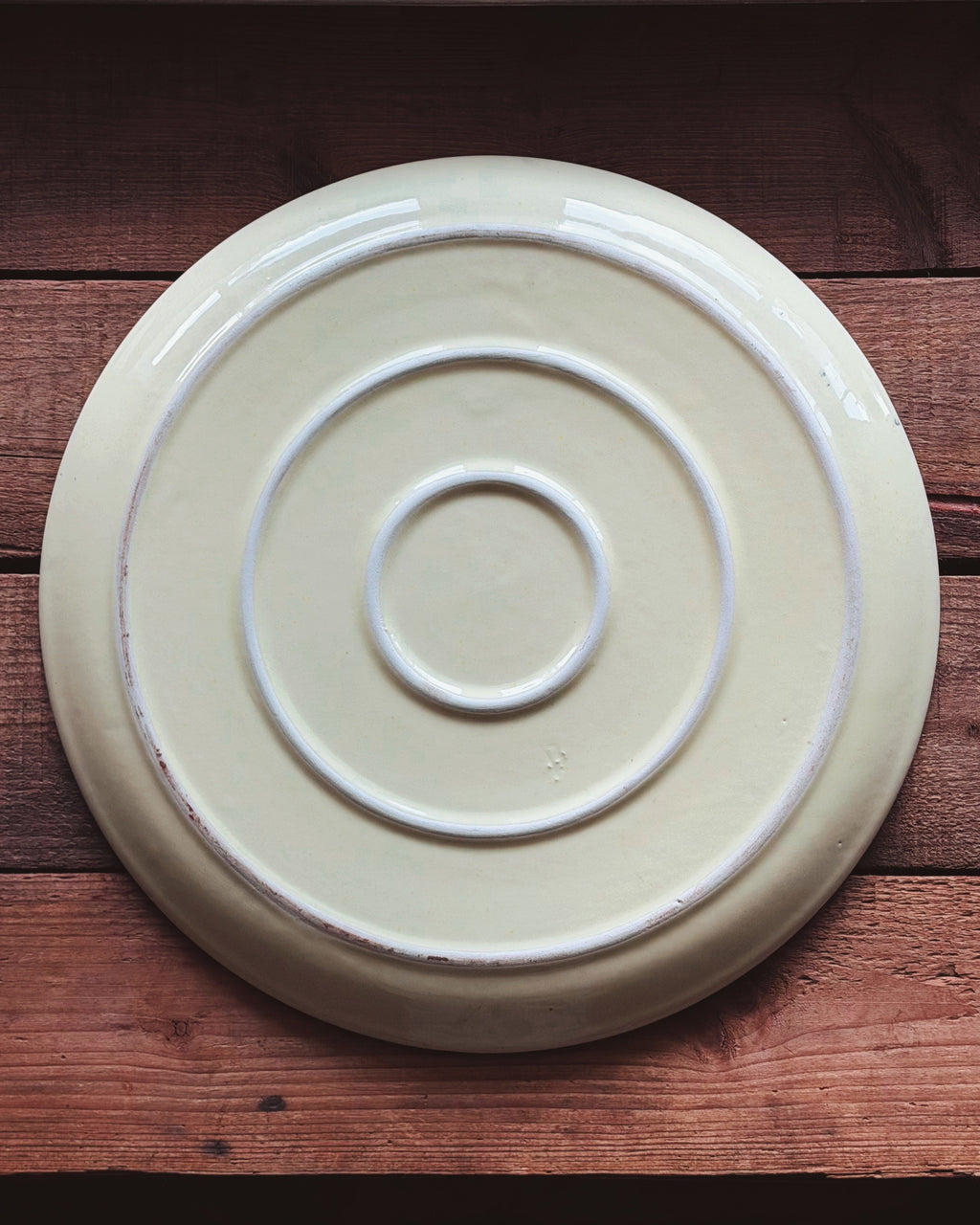 A round ceramic plate with concentric circular ridges, shown from above on a rustic wooden surface. The plate has a warm cream or off-white glaze with a subtle distressed finish along the edges.