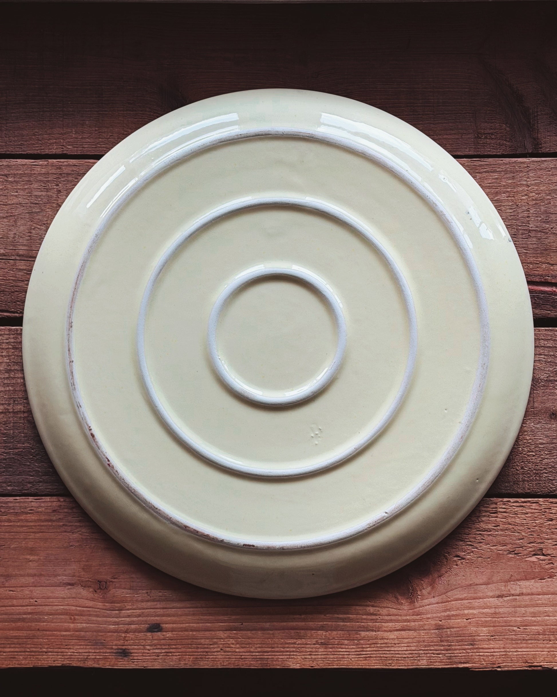 A round ceramic plate with concentric circular ridges, shown from above on a rustic wooden surface. The plate has a warm cream or off-white glaze with a subtle distressed finish along the edges.