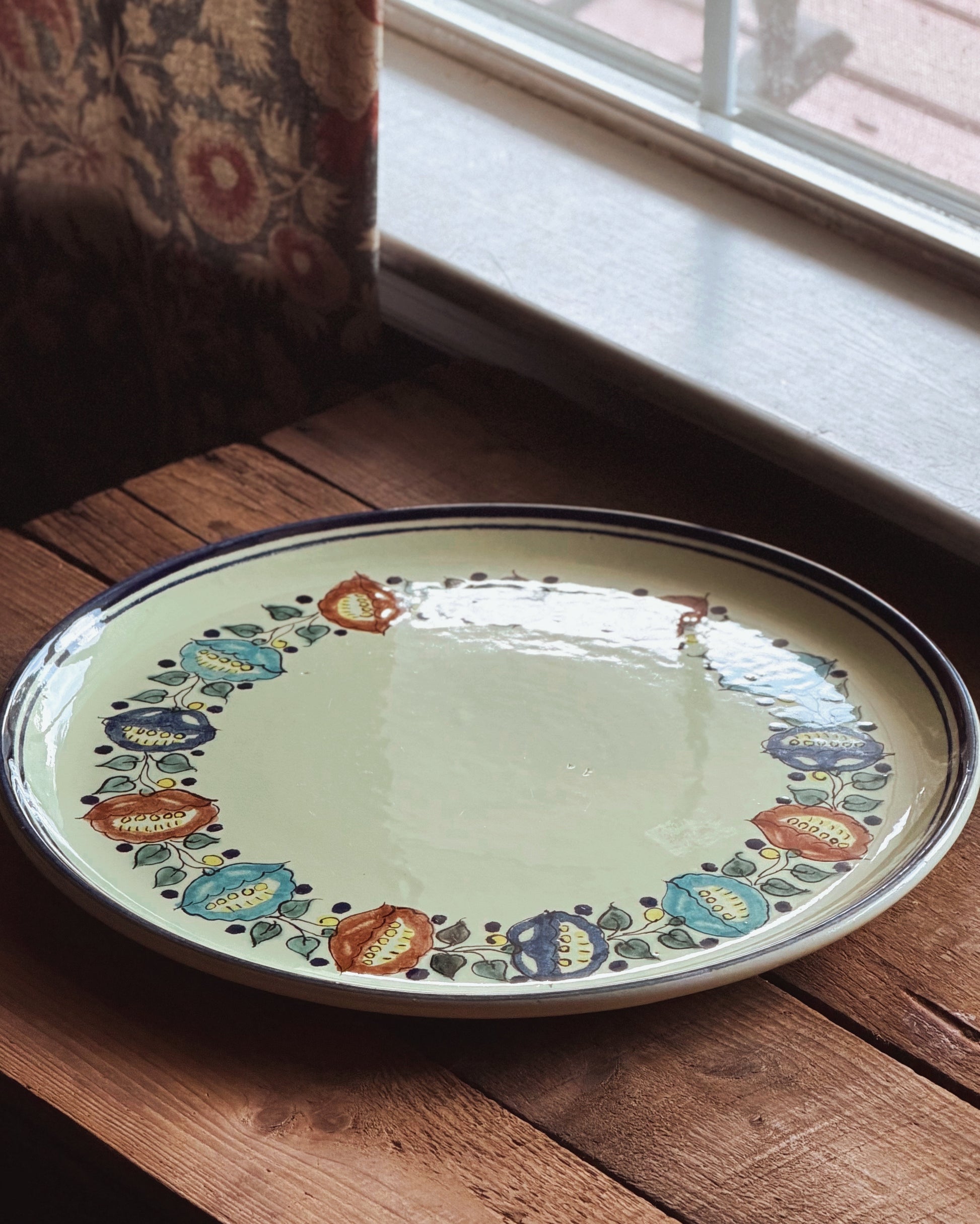 A round ceramic plate with a pale yellow center and a hand-painted floral border in blue, orange, and green, resting on a rustic wooden table near a window.