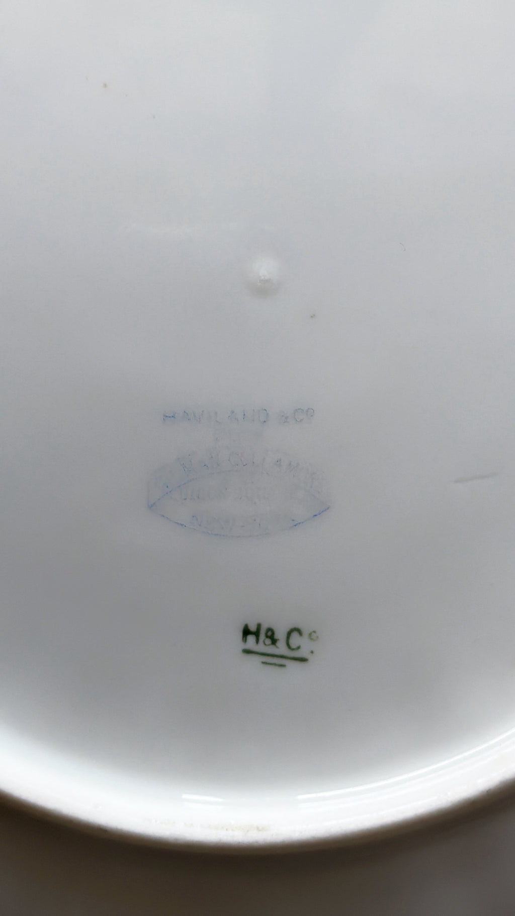 Close-up of the inside bottom of a white ceramic dish or bowl, showing faint blue and green printed maker marks and a small 'H&C' mark.