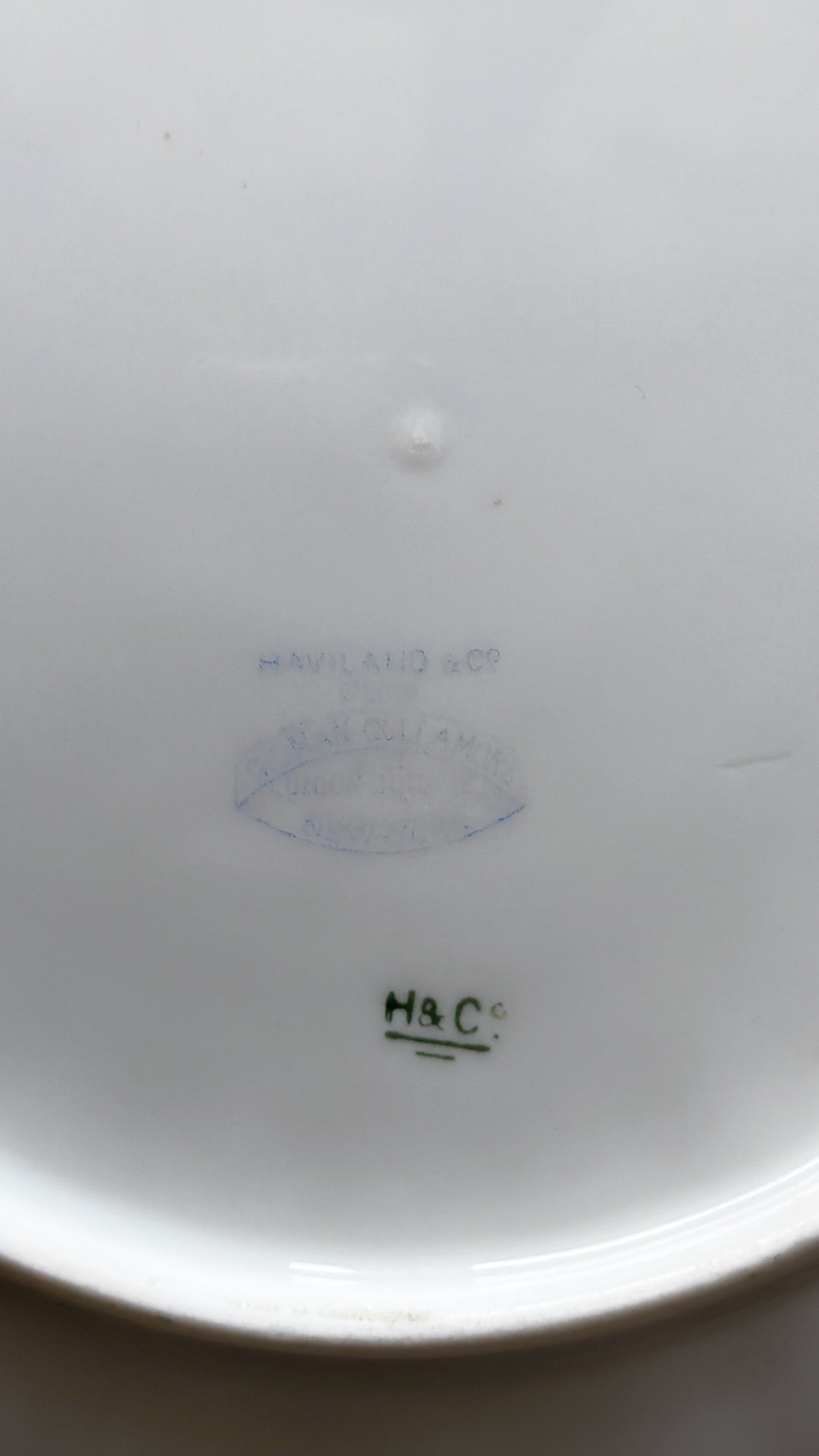 Close-up of the inside bottom of a white ceramic dish or bowl, showing faint blue and green printed maker marks and a small 'H&C' mark.