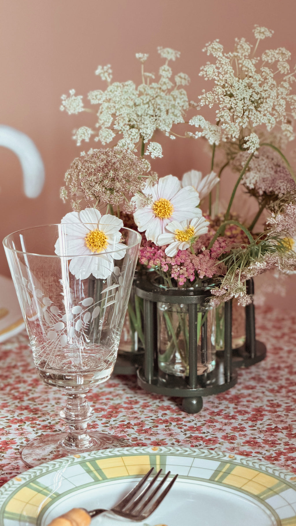 A styled tabletop floral arrangement with white daisy-like flowers, pink blossoms, and delicate greenery in a Vintage Two’s Company Triple Bud Vase Centerpiece, alongside a clear etched glass goblet and a patterned plate on a matching tablecloth against a soft blush background.