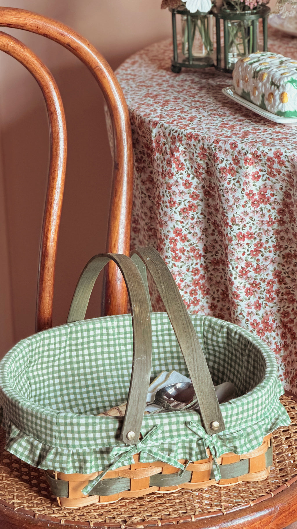 A woven basket with a green and white gingham fabric liner and olive green handles, resting on a wooden chair in a vintage-style room with a floral tablecloth.