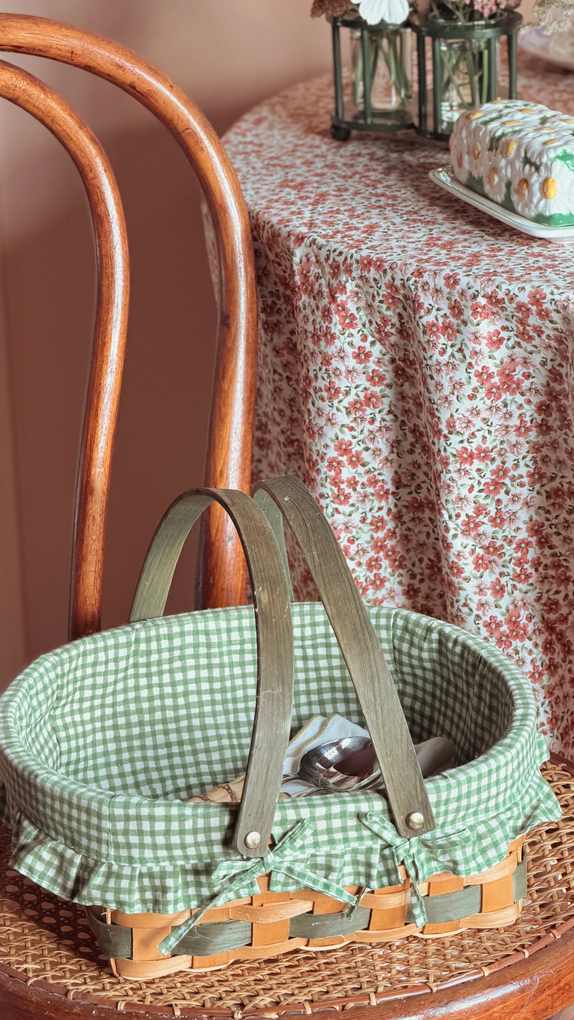 A woven basket with a green and white gingham fabric liner and olive green handles, resting on a wooden chair in a vintage-style room with a floral tablecloth.