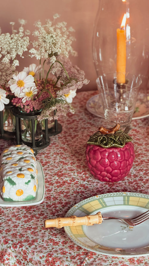 A styled tabletop scene with a floral tablecloth, a plate with a fork, a Vintage Ceramic Grape Jam Jar with Lid & Ladle, flowers in vases, and a lit candle in a glass holder.