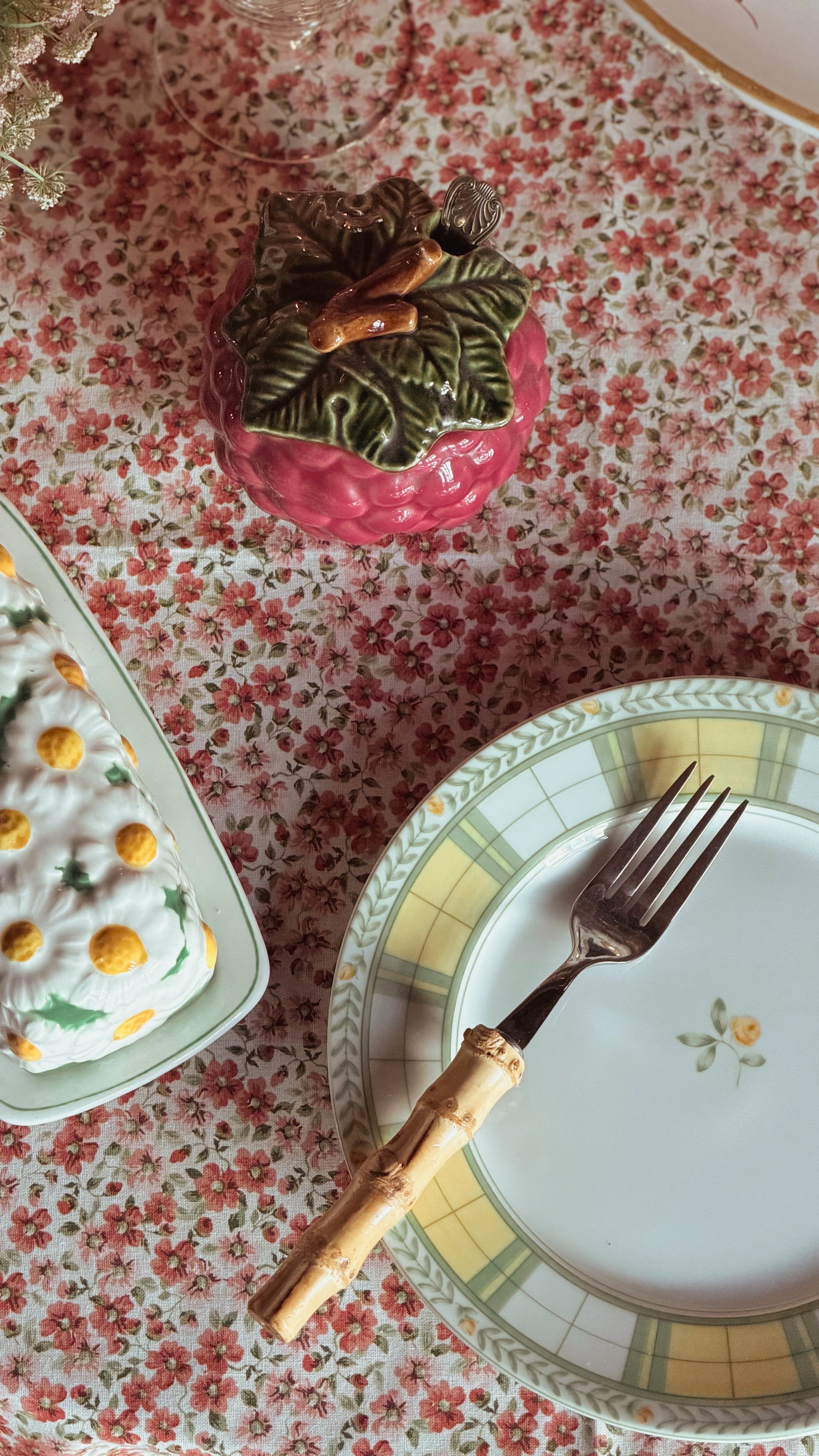 A styled tabletop scene with a white plate featuring a yellow and green checkered rim, a fork with a bamboo handle, a pink decorative container, and a floral-patterned tablecloth.
