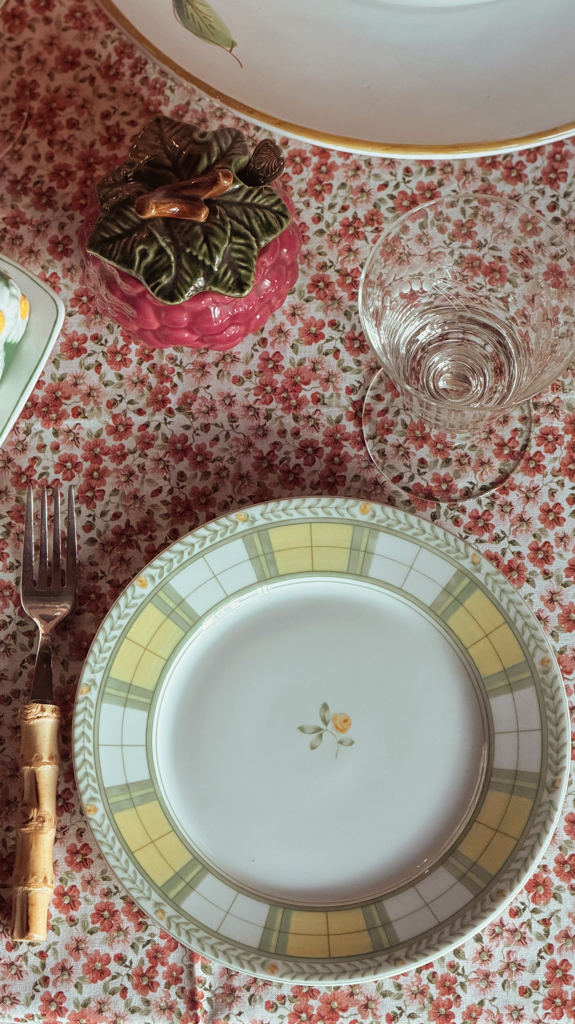 A top-down view of a table setting featuring a Vintage Mikasa English Rose Salad Plate with a yellow and green plaid border and a small floral motif in the center, placed on a red and white floral tablecloth next to a fork.