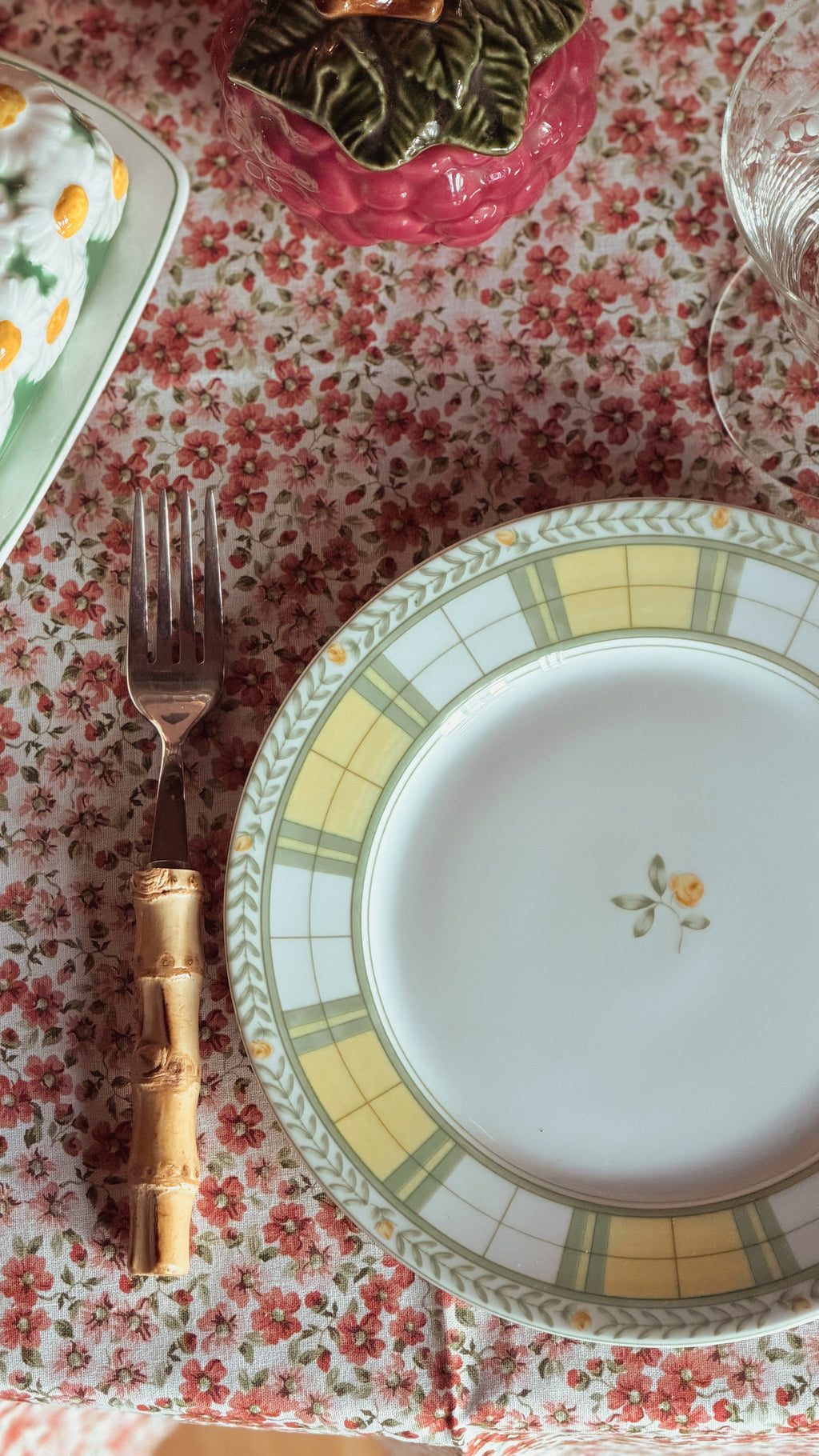 A close-up of a Vintage Mikasa English Rose Salad Plate with a yellow and green checkered border and a small floral motif in the center, placed on a red and white floral tablecloth next to a fork with a bamboo handle.