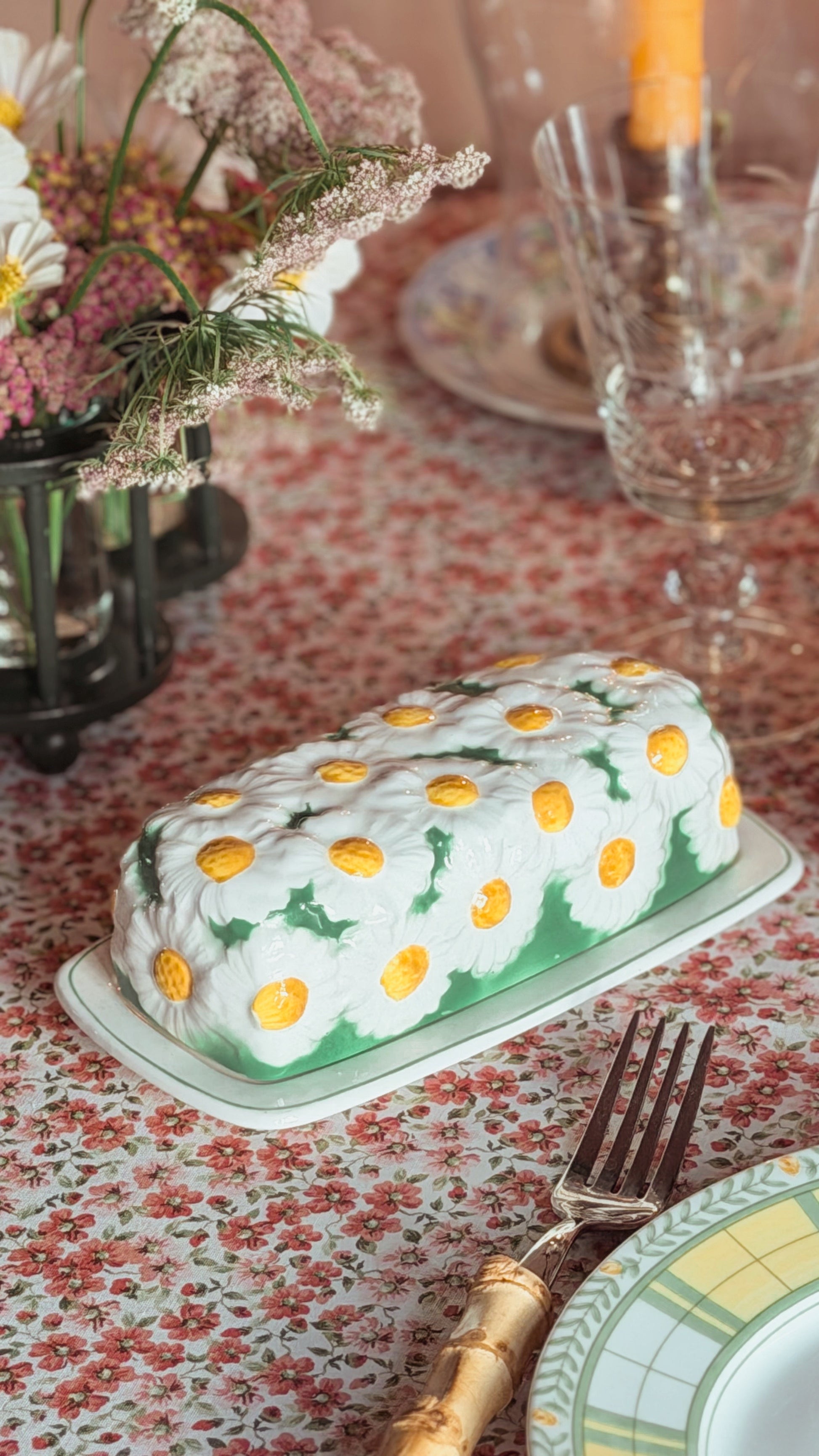 A Lefton Daisytime Ceramic Butter Dish featuring a white daisy pattern with yellow centers and green leaves, set on a floral tablecloth with a fork and glassware nearby.