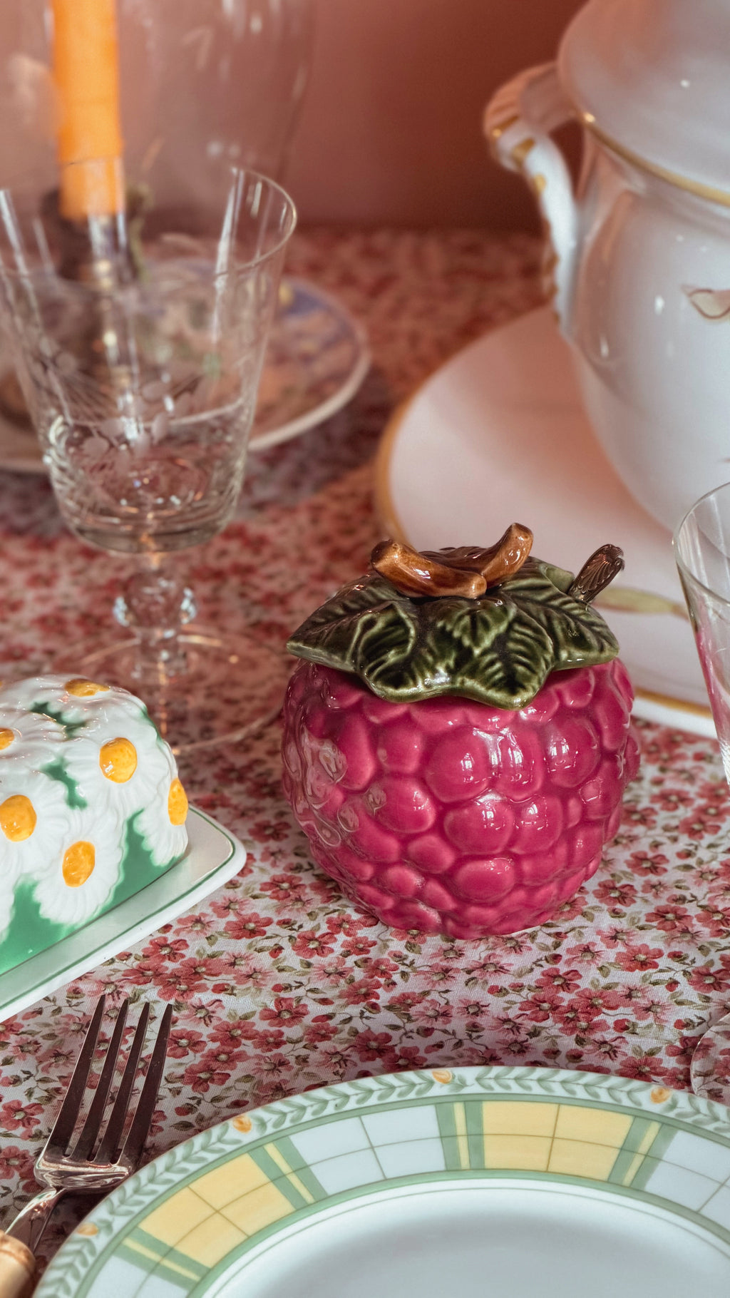 A pink raspberry-shaped ceramic container with a green leafy top and brown stem, styled on a floral tablecloth with plates, glasses, and cutlery in a tabletop setting.