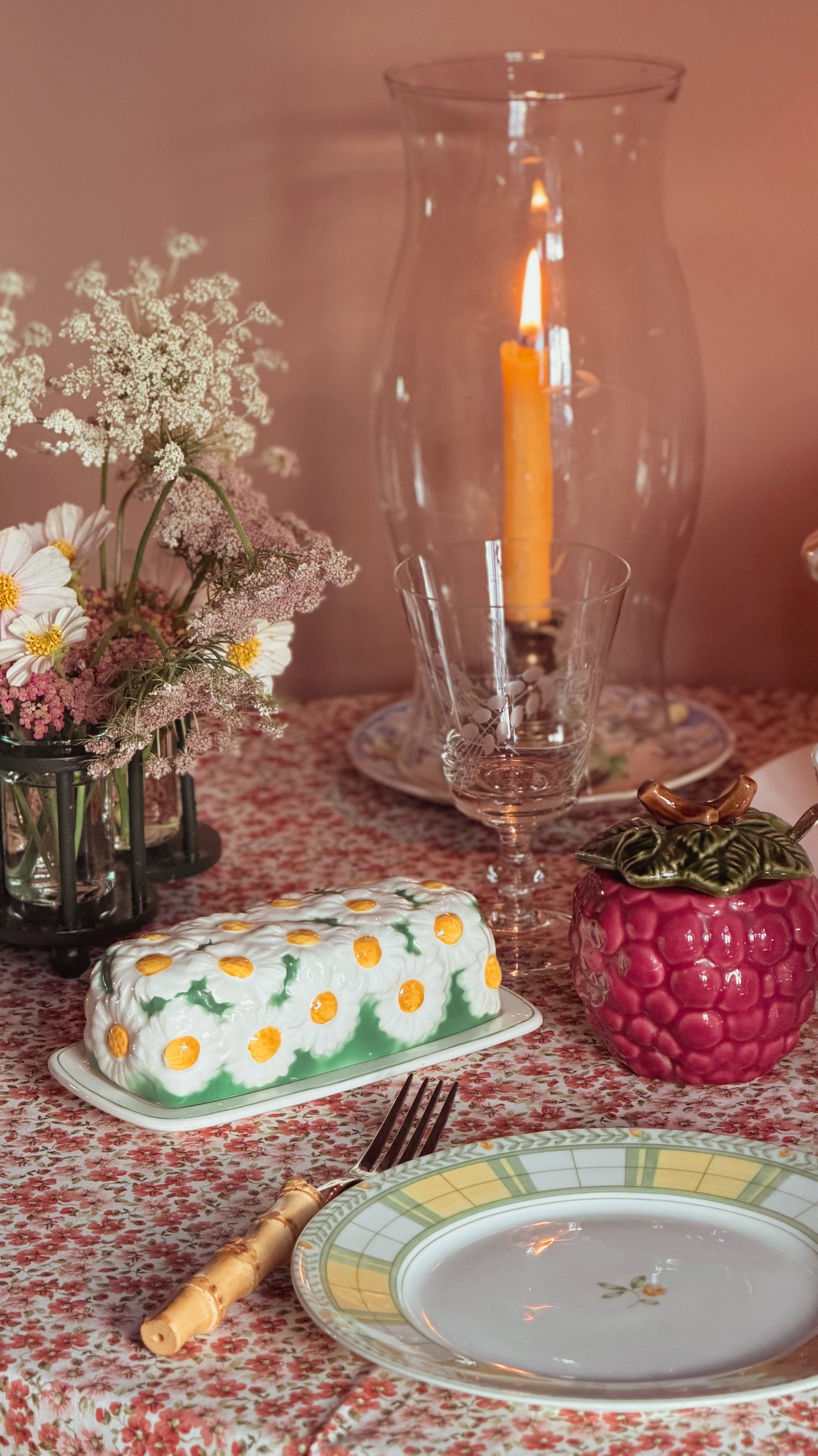 A styled tabletop scene with a butter dish covered in white daisy flowers with yellow centers, set on a red floral tablecloth beside a plate, candle holder with a lit orange candle, and a pink textured container.