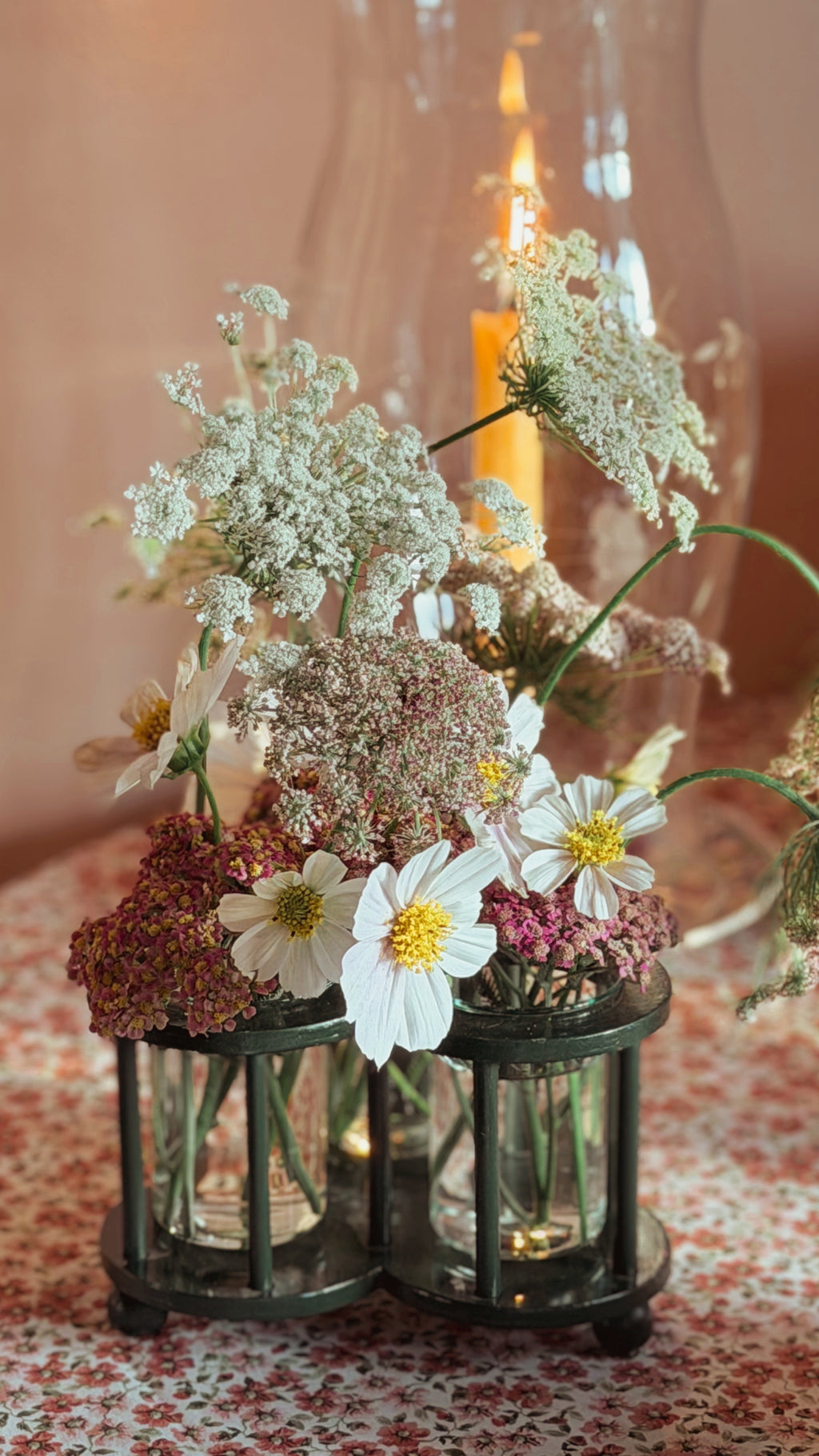 A rustic floral centerpiece arrangement in two small dark metal lantern-style holders, featuring white daisies, baby's breath, and pink wildflowers, with a blurred lit candle in the background.