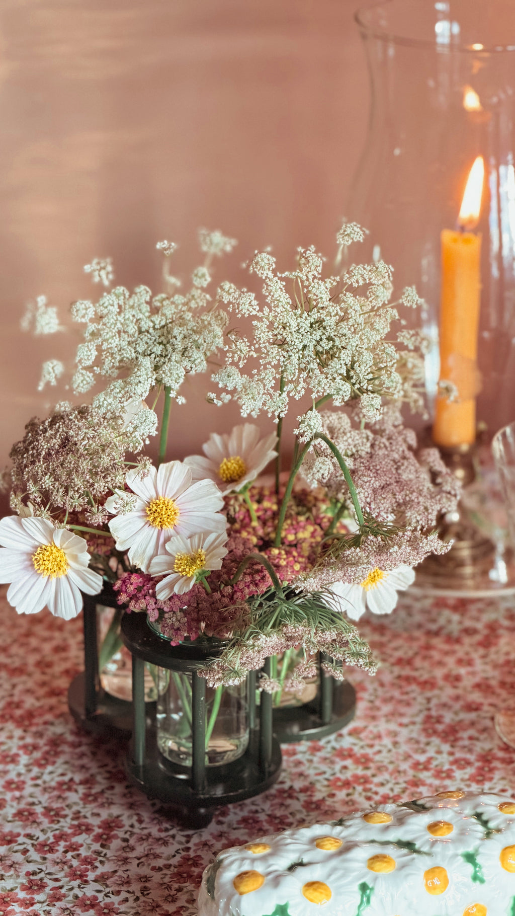 A rustic floral centerpiece in a dark glass vase, featuring white daisies, wildflowers, and baby's breath on a patterned tablecloth with a warm, candlelit background.