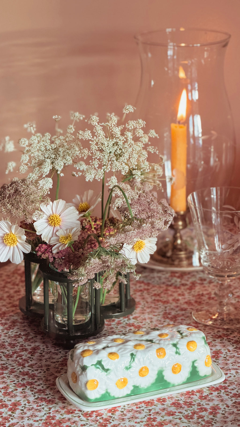 A butter dish decorated with a white daisy pattern and yellow centers, set on a floral tablecloth next to a Vintage Two’s Company Triple Bud Vase Centerpiece of white flowers and a lit candle in a tall glass holder.