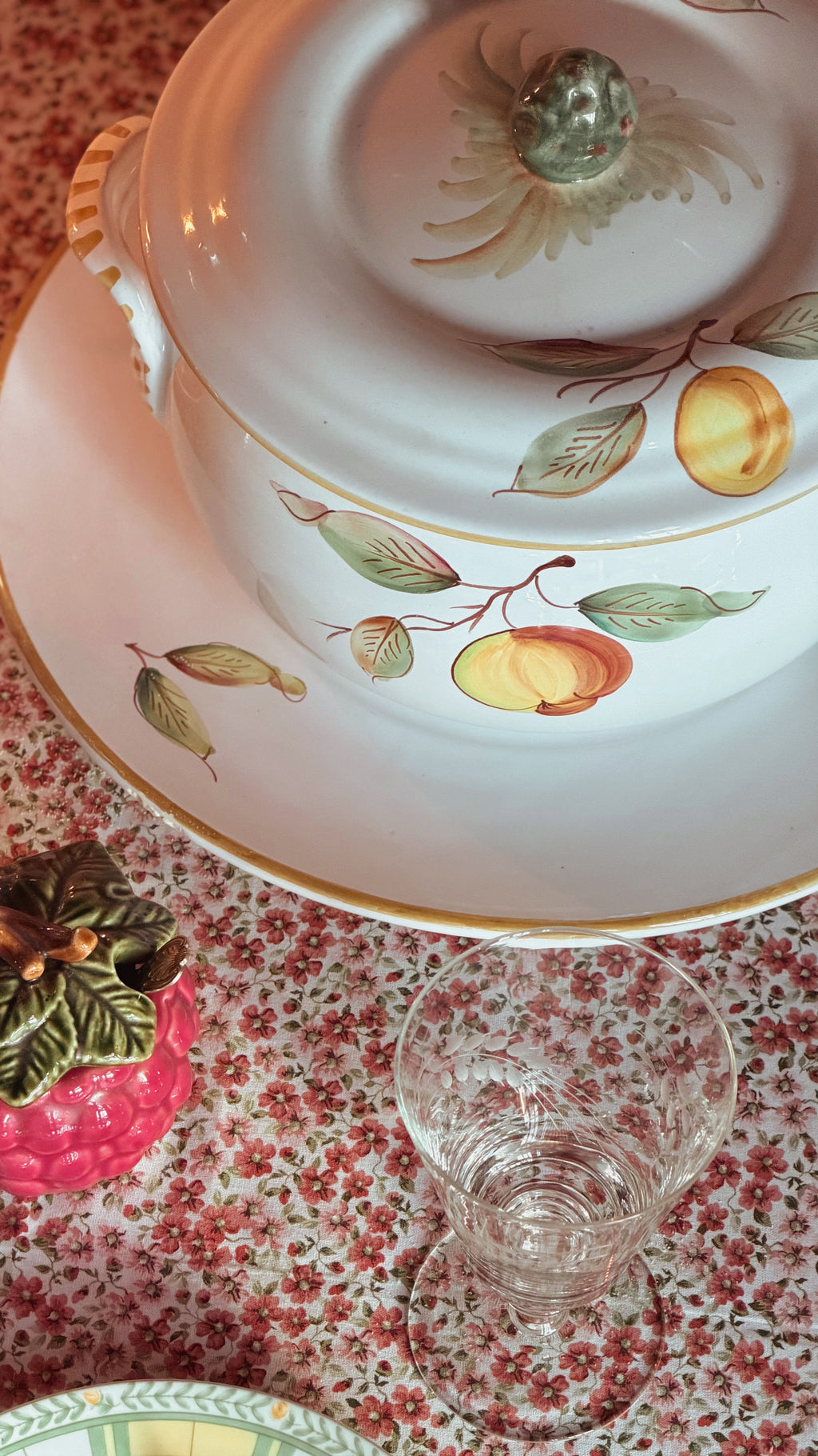 A Vintage Ceramiche Fontana Siena CF62 Hand Painted Italian Tureen with Ladle & Underplate, shown on a floral tablecloth with a small clear glass and a pink decorative object nearby.