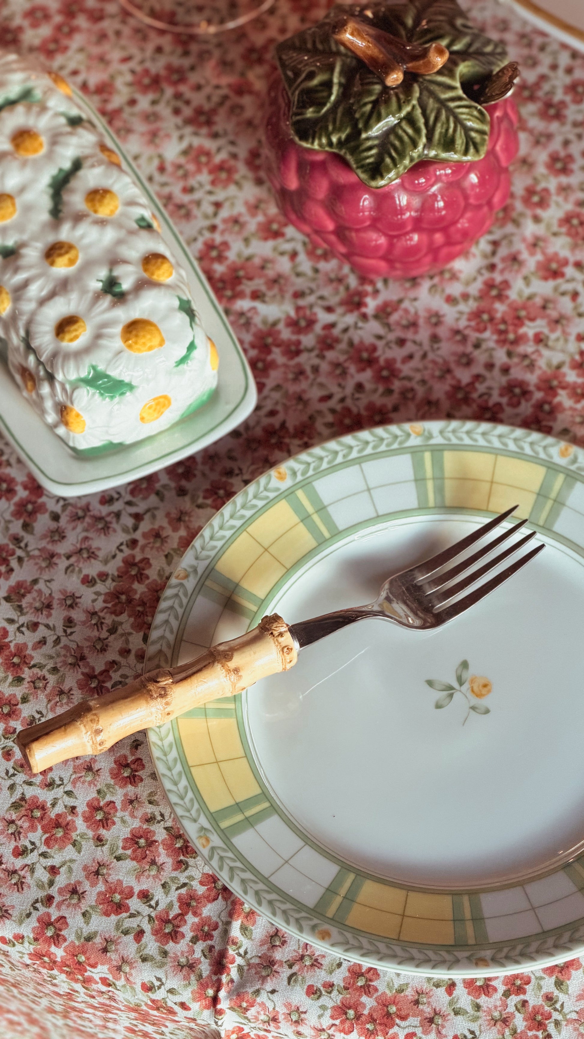 A close-up of a Vintage Mikasa English Rose Salad Plate with a bamboo-handled fork resting on it, set on a red and white floral tablecloth. In the background, there is a pink raspberry-shaped container and a white rectangular dish with yellow and green floral details.