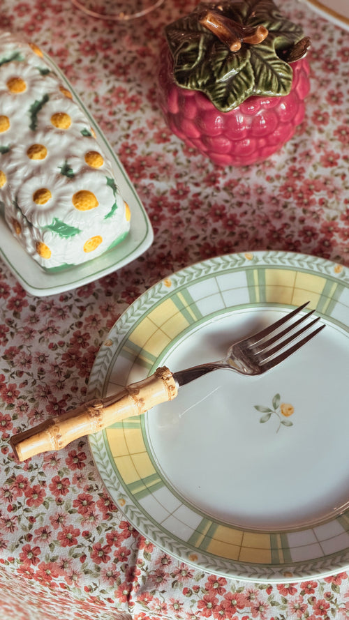 A close-up of a Vintage Mikasa English Rose Salad Plate with a bamboo-handled fork resting on it, set on a red and white floral tablecloth. In the background, there is a pink raspberry-shaped container and a white rectangular dish with yellow and green floral details.