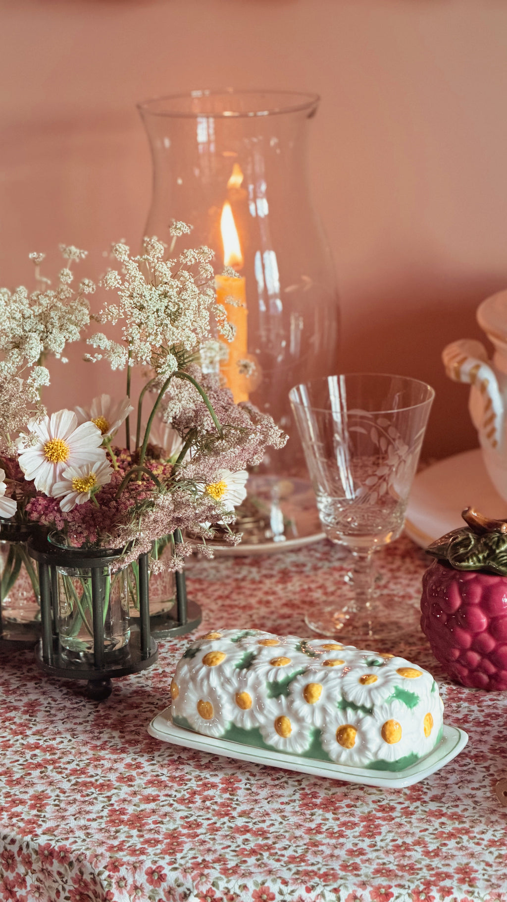 A butter dish decorated with white daisy flowers and yellow centers, resting on a floral tablecloth next to a glass vase with dried flowers and a lit candle.