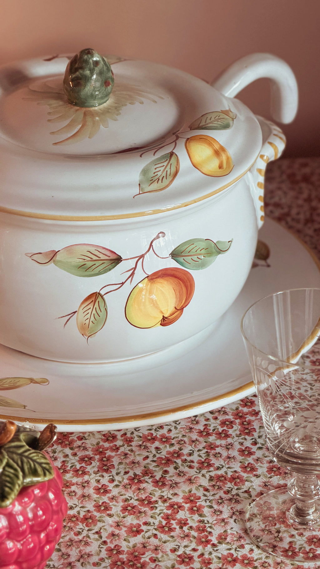 A white porcelain lidded Vintage Ceramiche Fontana Siena CF62 Hand Painted Italian Tureen with Ladle & Underplate decorated with painted fruit and leaf motifs, shown on a matching saucer over a floral tablecloth, with a glass and a decorative fruit prop nearby.