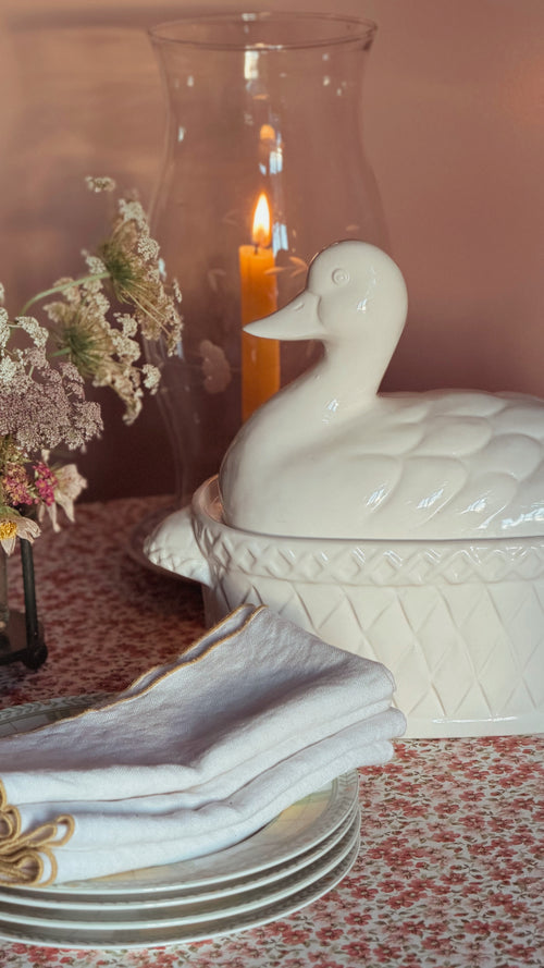 A white ceramic duck-shaped lidded tureen sits on a patterned tablecloth beside folded white napkins, plates, dried flowers, and a lit candle in a glass holder.