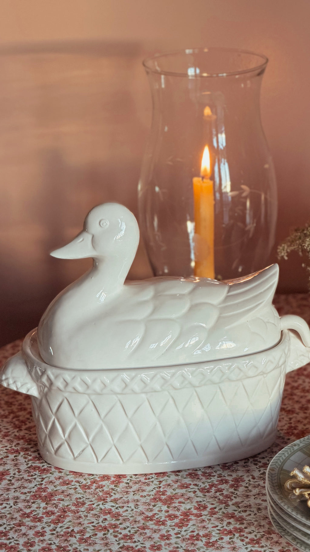 A white ceramic duck-shaped lidded dish with a textured diamond-patterned base, set on a floral tablecloth with a lit candle in a glass holder behind it.