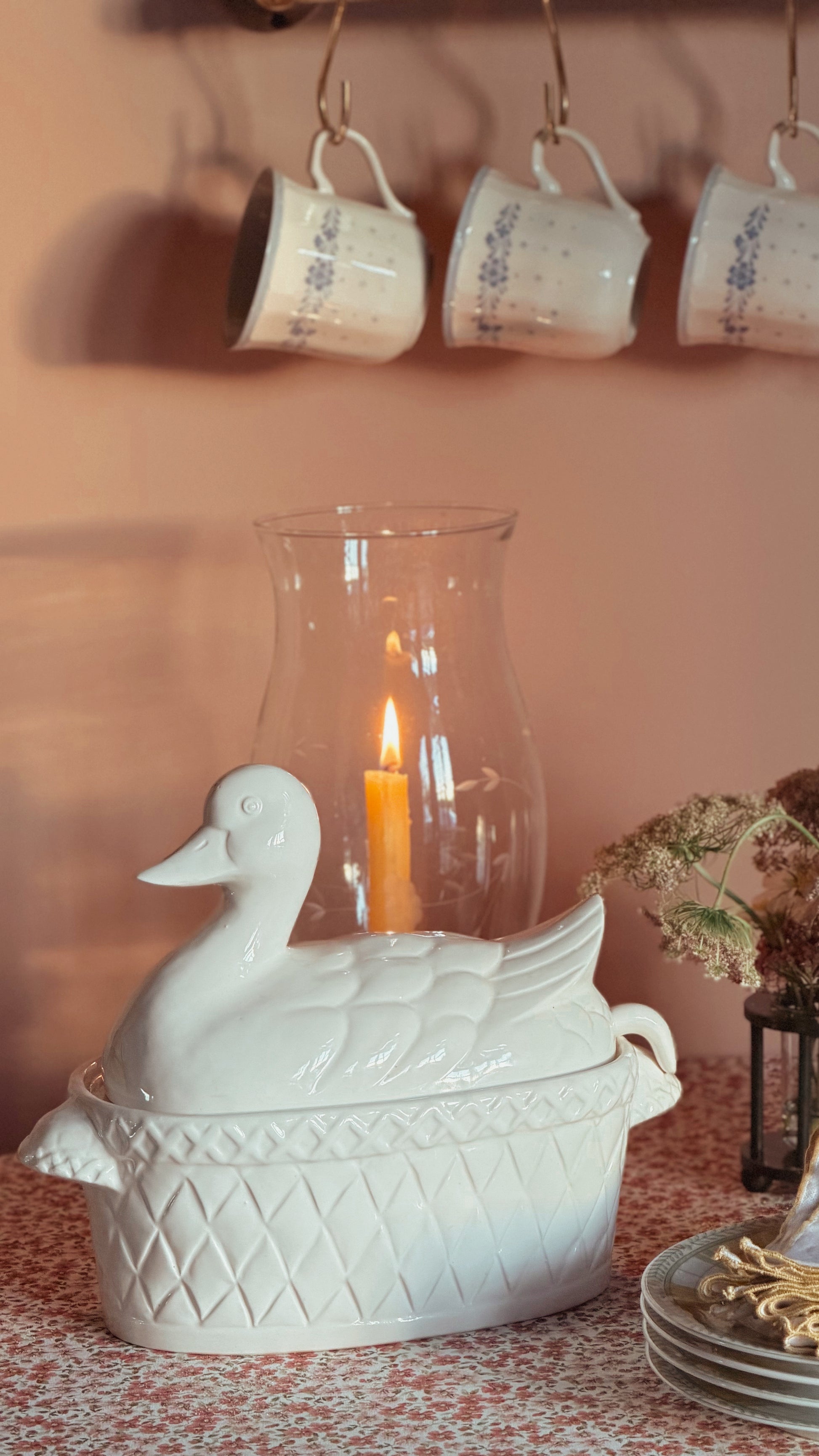 A Vintage White Ceramic Duck Tureen with Ladle on a patterned table surface, with a lit orange candle inside and a clear glass vase behind it. A peach-colored wall and three hanging white mugs are visible in the background.