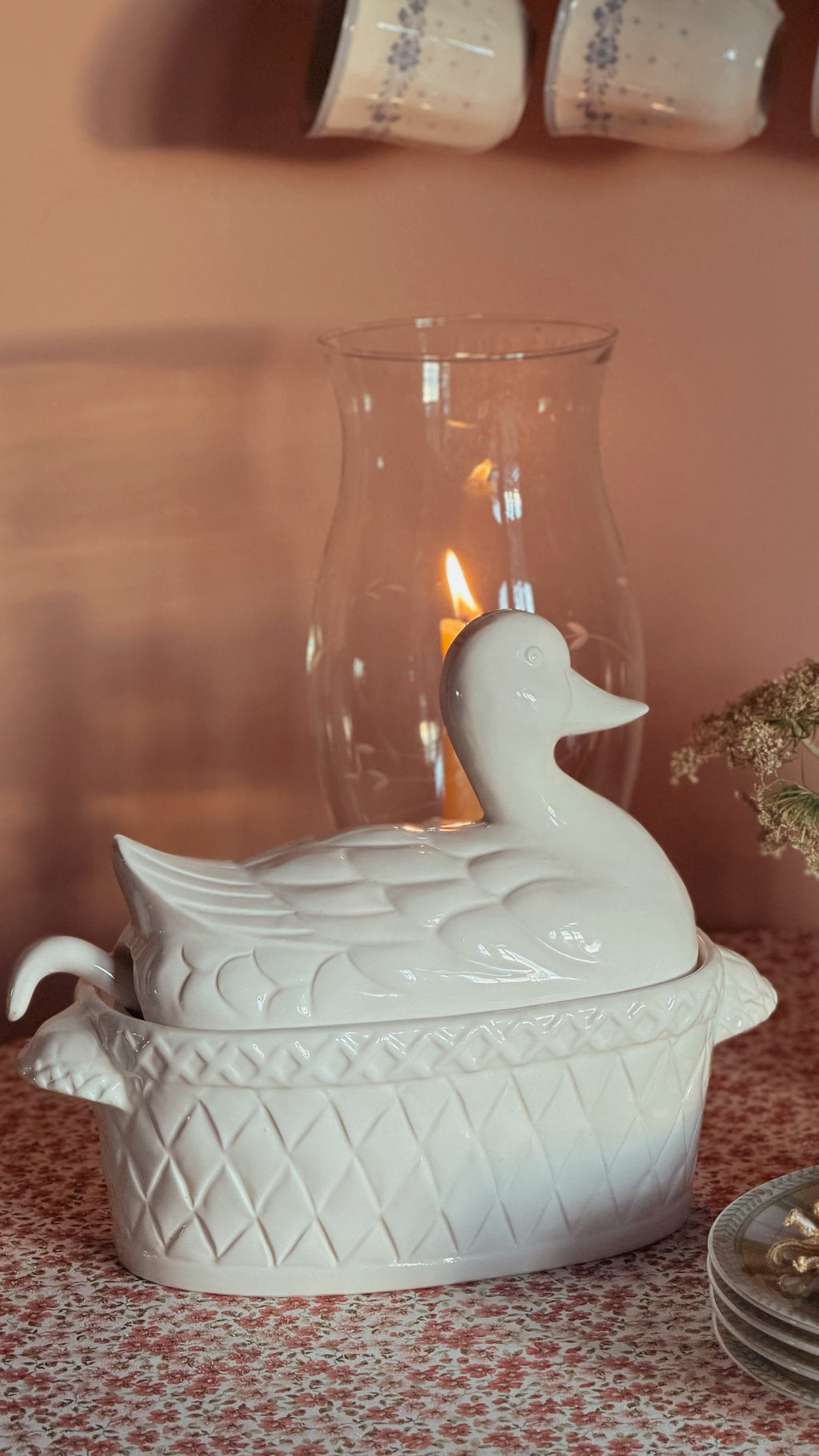 A white ceramic duck-shaped lidded dish or tureen sits on a patterned tablecloth in a styled interior setting, with a glass candle holder and warm light in the background.