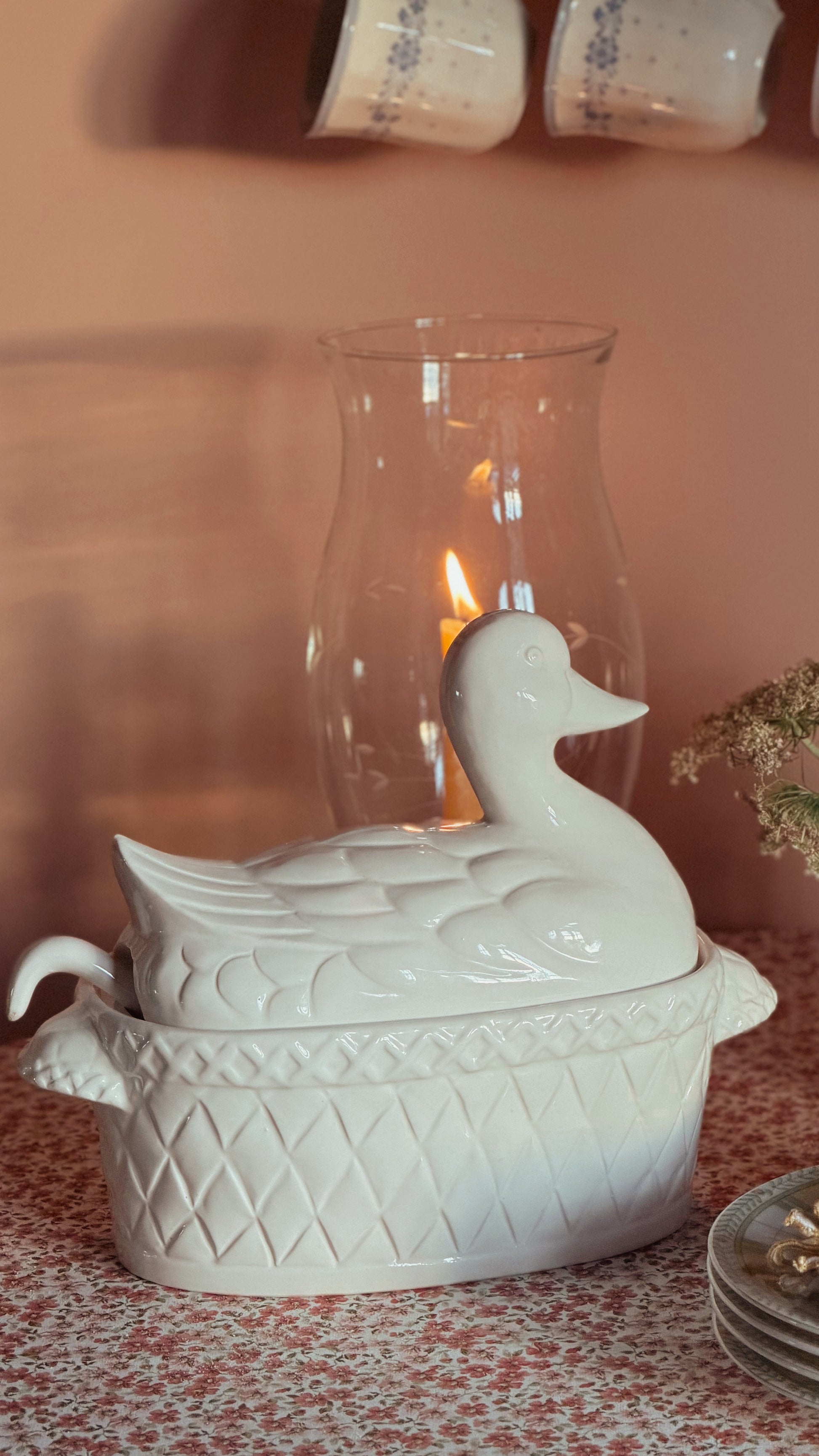 A white ceramic duck-shaped lidded dish or tureen sits on a patterned tablecloth in a styled interior setting, with a glass candle holder and warm light in the background.