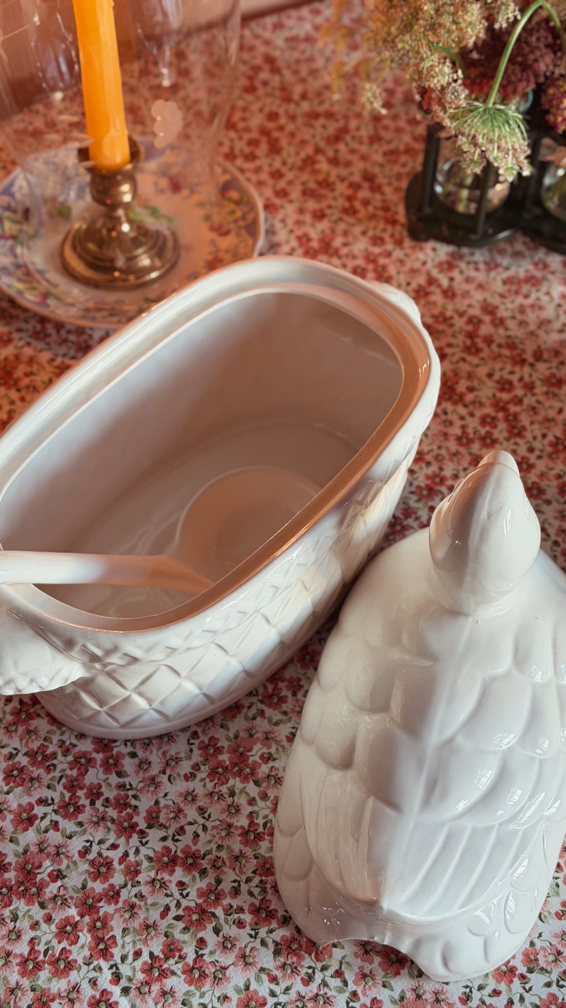 A Vintage White Ceramic Duck Tureen with Ladle, shown open with ladle inside, set on a floral tablecloth with a candle and flowers in the background.