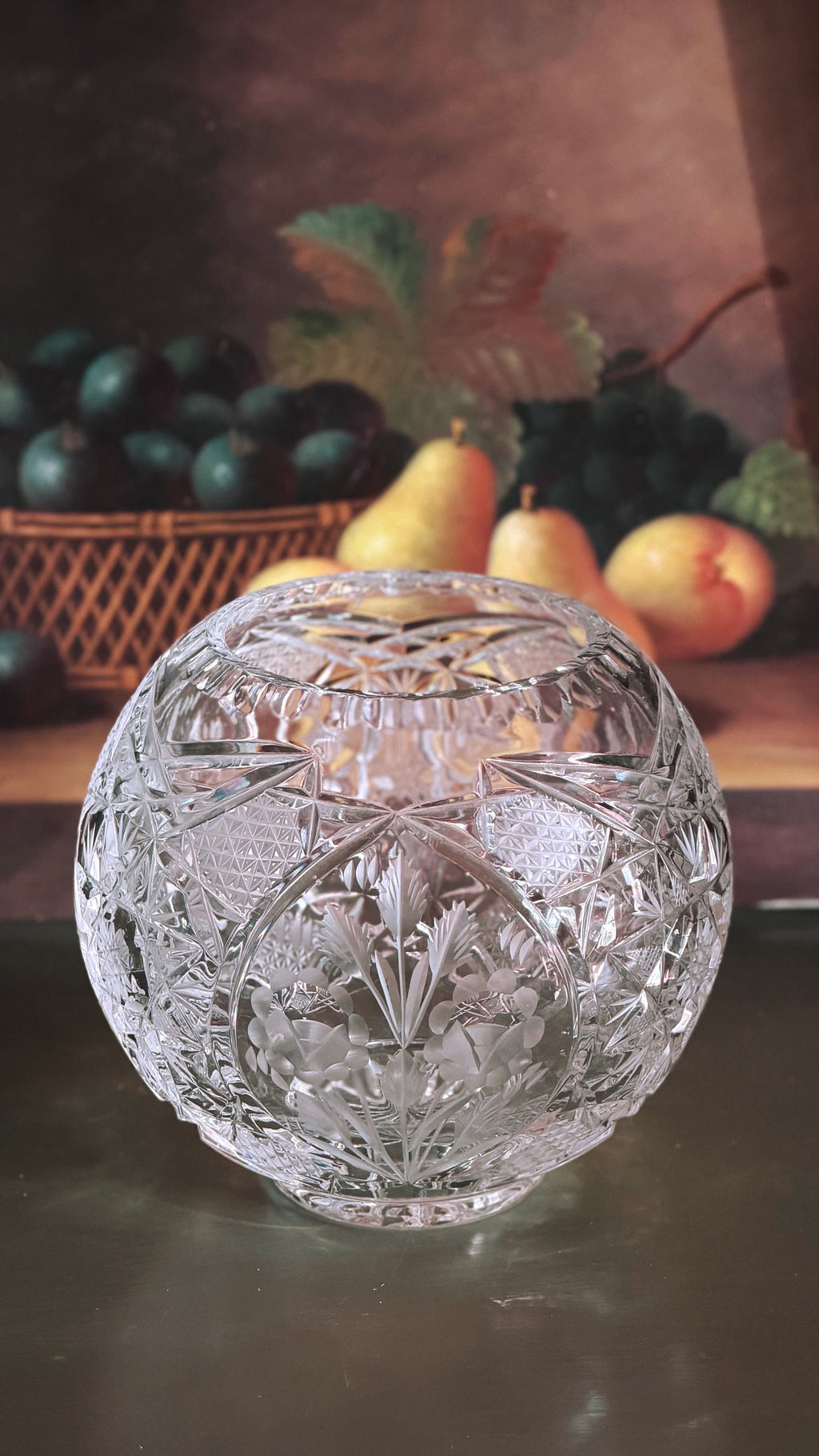 A round, clear Vintage Etched Crystal Rose Bowl Vase with intricate floral and geometric patterns, set against a dark, rustic background with blurred fruit.