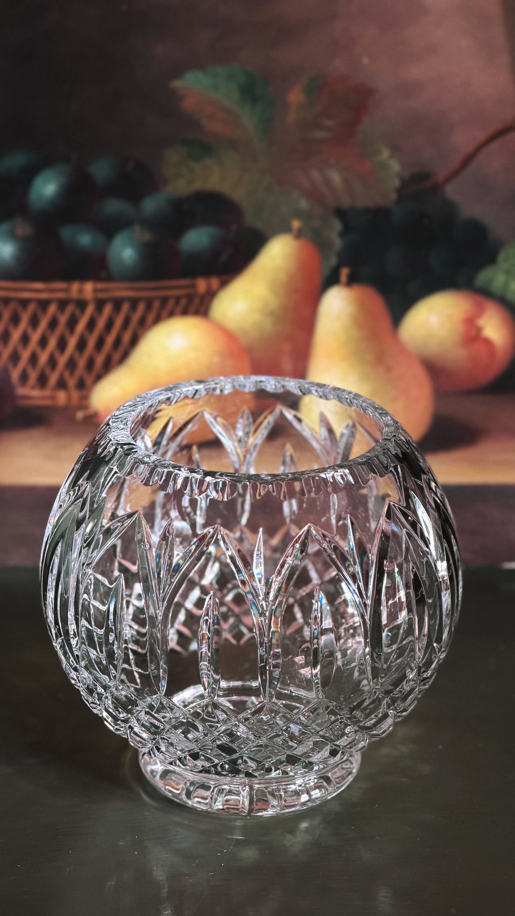 A clear cut-crystal fishbowl vase with intricate geometric facets, displayed on a dark reflective surface with a blurred fruit still life background.