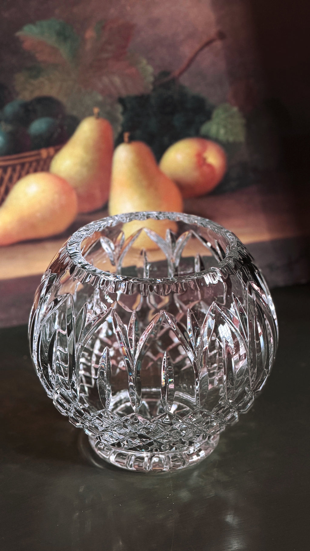 Vintage Crystal Rose Bowl Vase with an intricate geometric pattern, set on a dark reflective surface with a blurred fruit painting in the background.