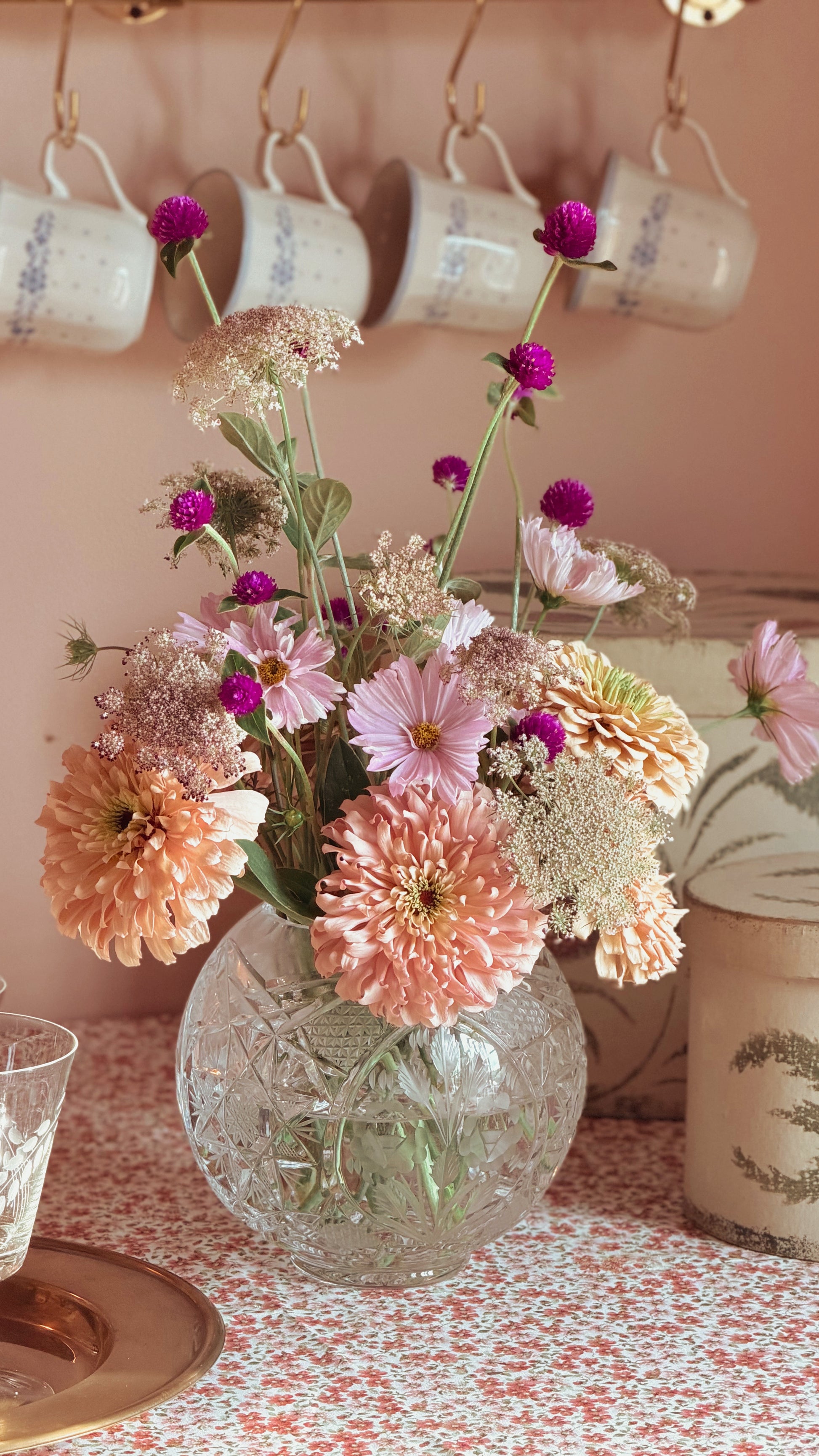 A floral arrangement of pink, peach, and purple flowers in a clear glass vase, set on a patterned tablecloth with hanging ceramic mugs and a candle in the background.