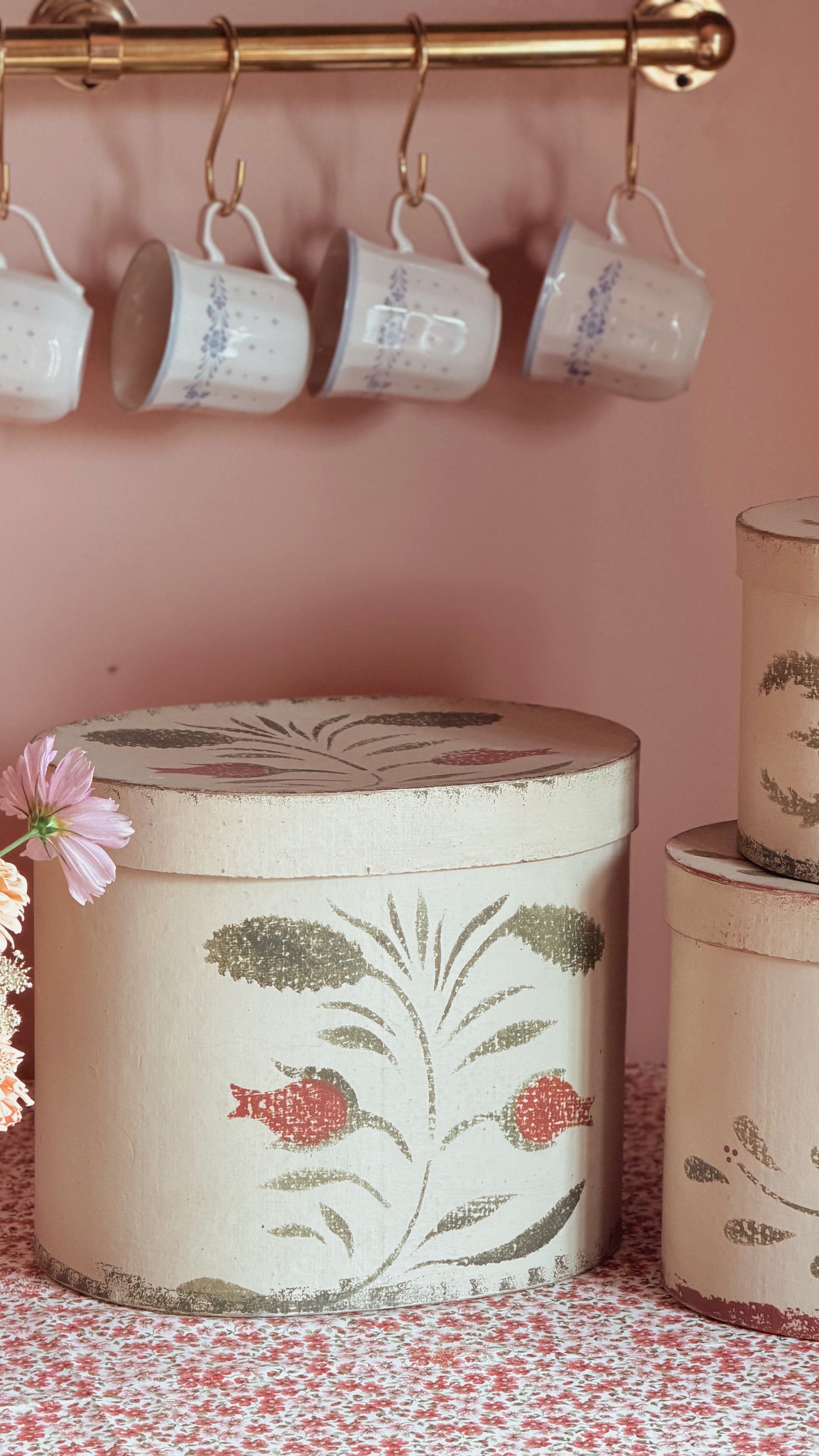 A set of vintage-style decorative storage boxes with a matching lid, featuring a botanical and fish pattern in muted green and red on a cream background. The boxes are styled on a pink floral tablecloth next to a pink flower, with matching white mugs hanging from a brass rod on a dusty pink wall in the background.
