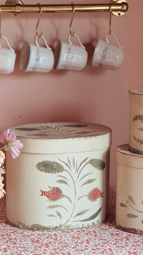 A set of vintage-style decorative storage boxes with a matching lid, featuring a botanical and fish pattern in muted green and red on a cream background. The boxes are styled on a pink floral tablecloth next to a pink flower, with matching white mugs hanging from a brass rod on a dusty pink wall in the background.