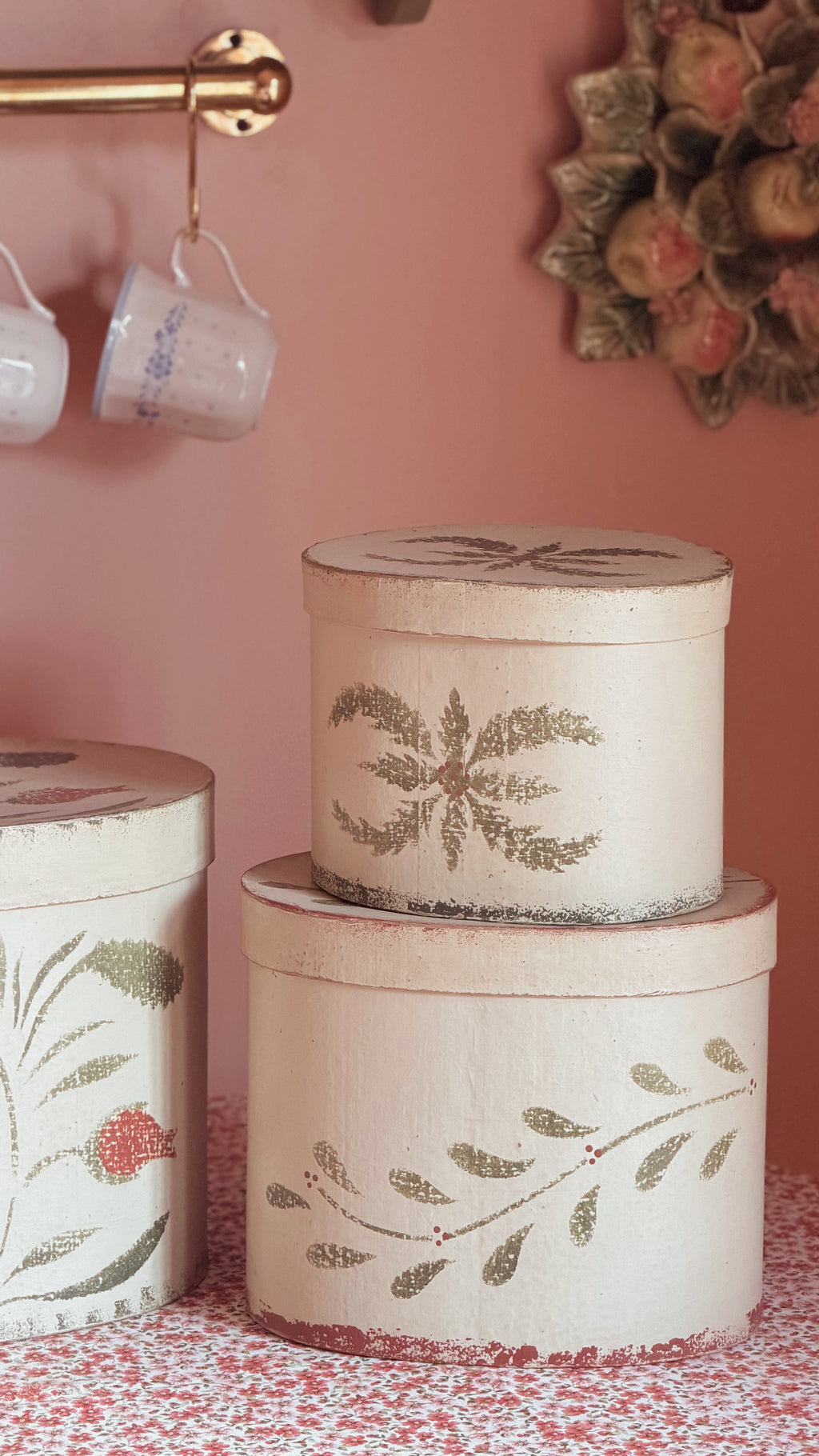 Three round decorative storage boxes stacked and displayed on a patterned surface against a pink wall. The boxes are off-white with distressed edges and painted botanical leaf motifs in muted green and brown tones.