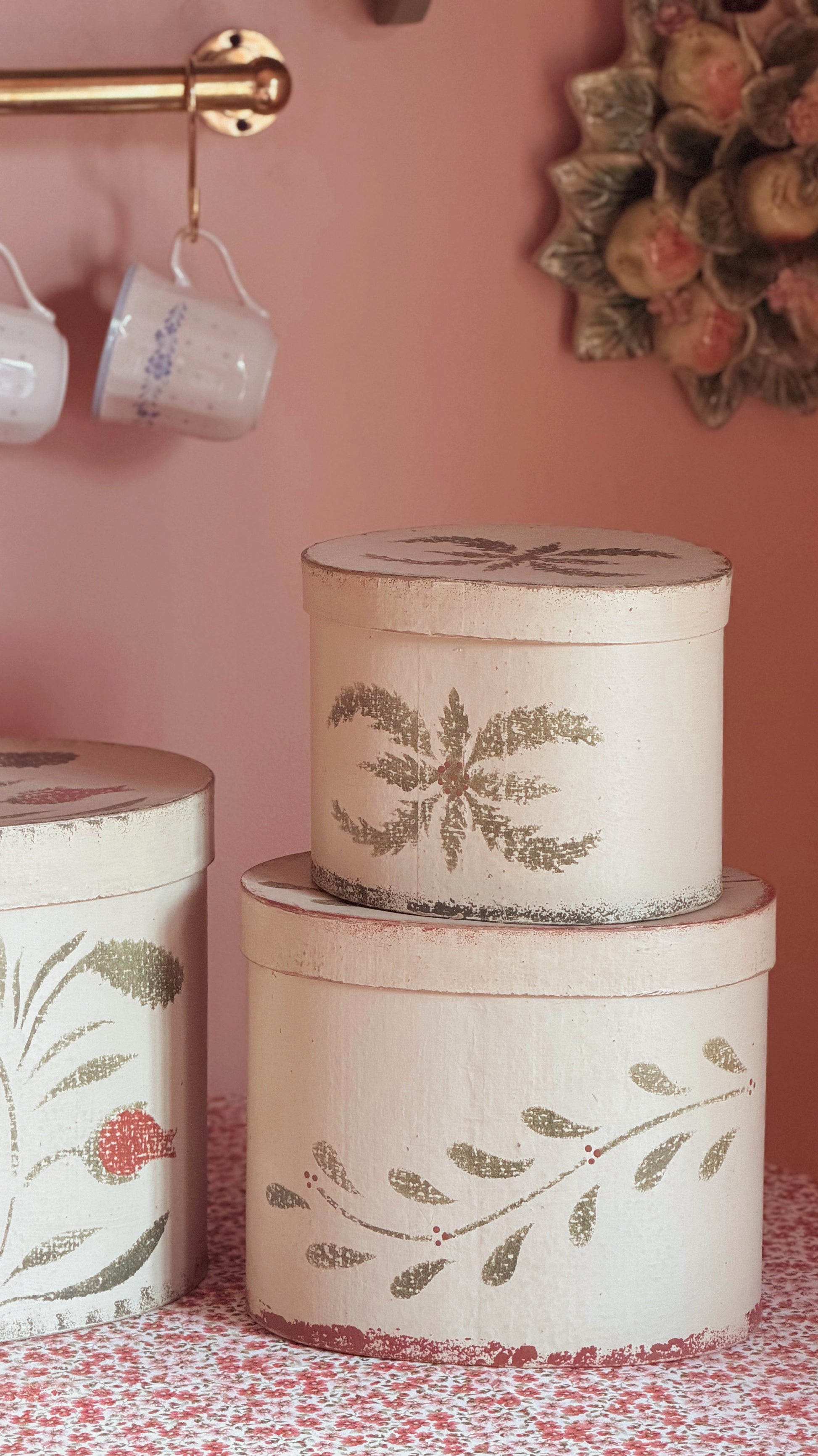Three round decorative storage boxes stacked and displayed on a patterned surface against a pink wall. The boxes are off-white with distressed edges and painted botanical leaf motifs in muted green and brown tones.