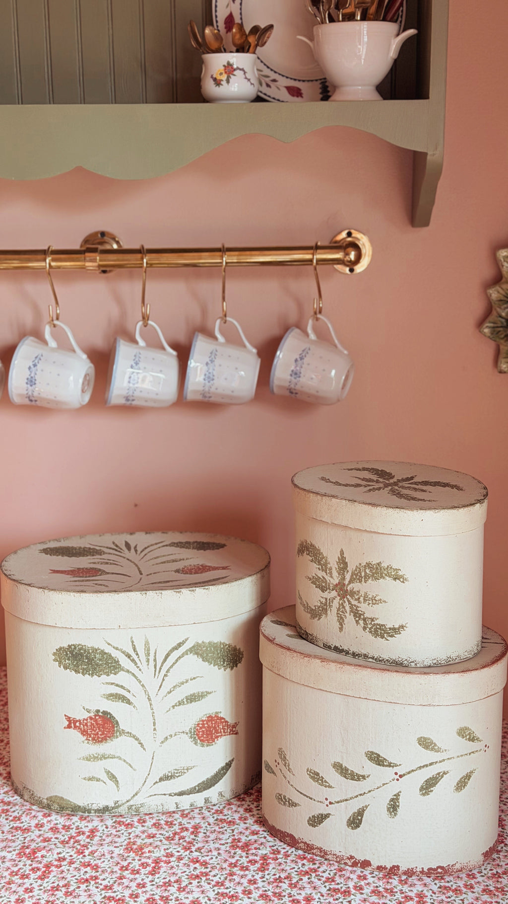 A styled interior scene with a pink wall, a brass hanging rod holding four white mugs, and three round decorative storage boxes with floral and leaf illustrations on a patterned surface.