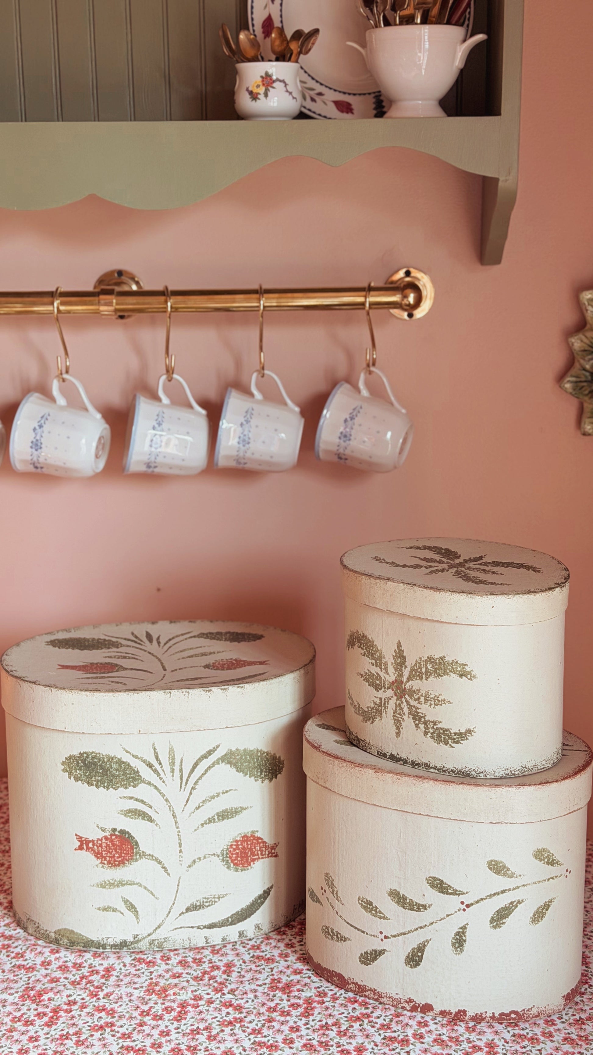 A styled interior scene with a pink wall, a brass hanging rod holding four white mugs, and three round decorative storage boxes with floral and leaf illustrations on a patterned surface.