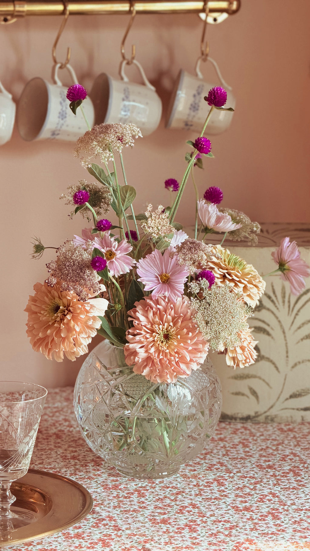A bouquet of mixed flowers in shades of pink, peach, and purple arranged in a Vintage Etched Crystal Rose Bowl Vase on a patterned tablecloth, with hanging white mugs on a wall in the background.